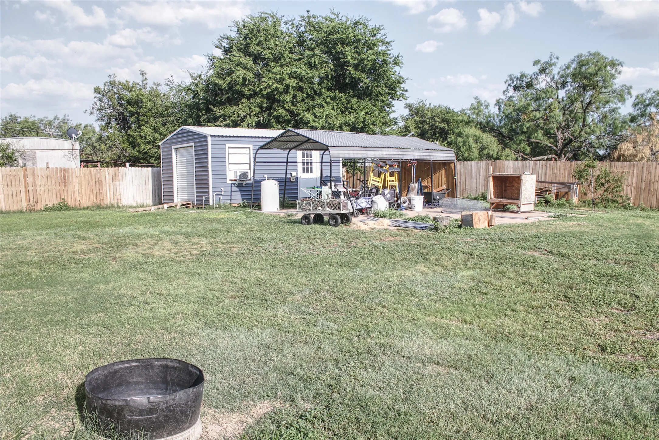 Expansive grass lawn featuring a wood privacy fence, a blue metal storage shed, and a detached covered carport