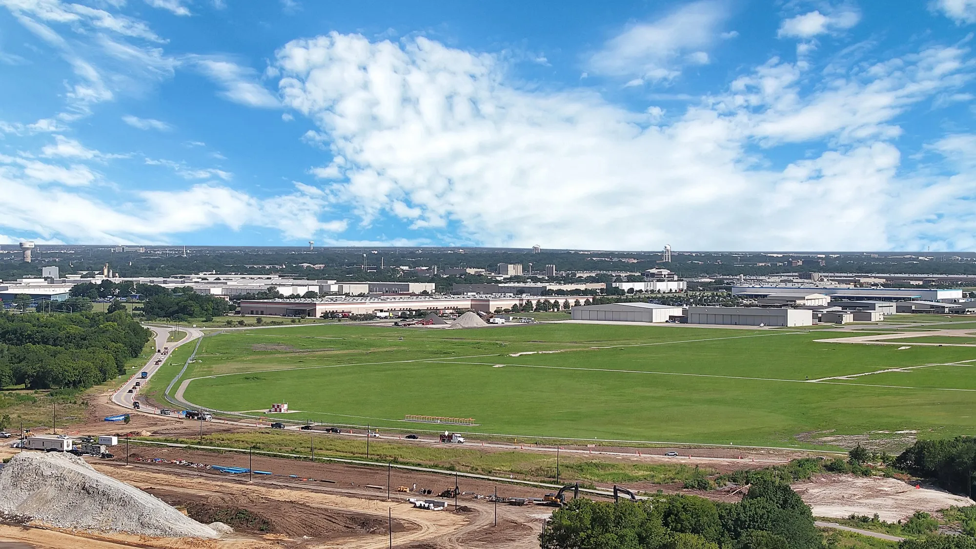Expansive green fields with distant industrial buildings under a blue sky with white clouds