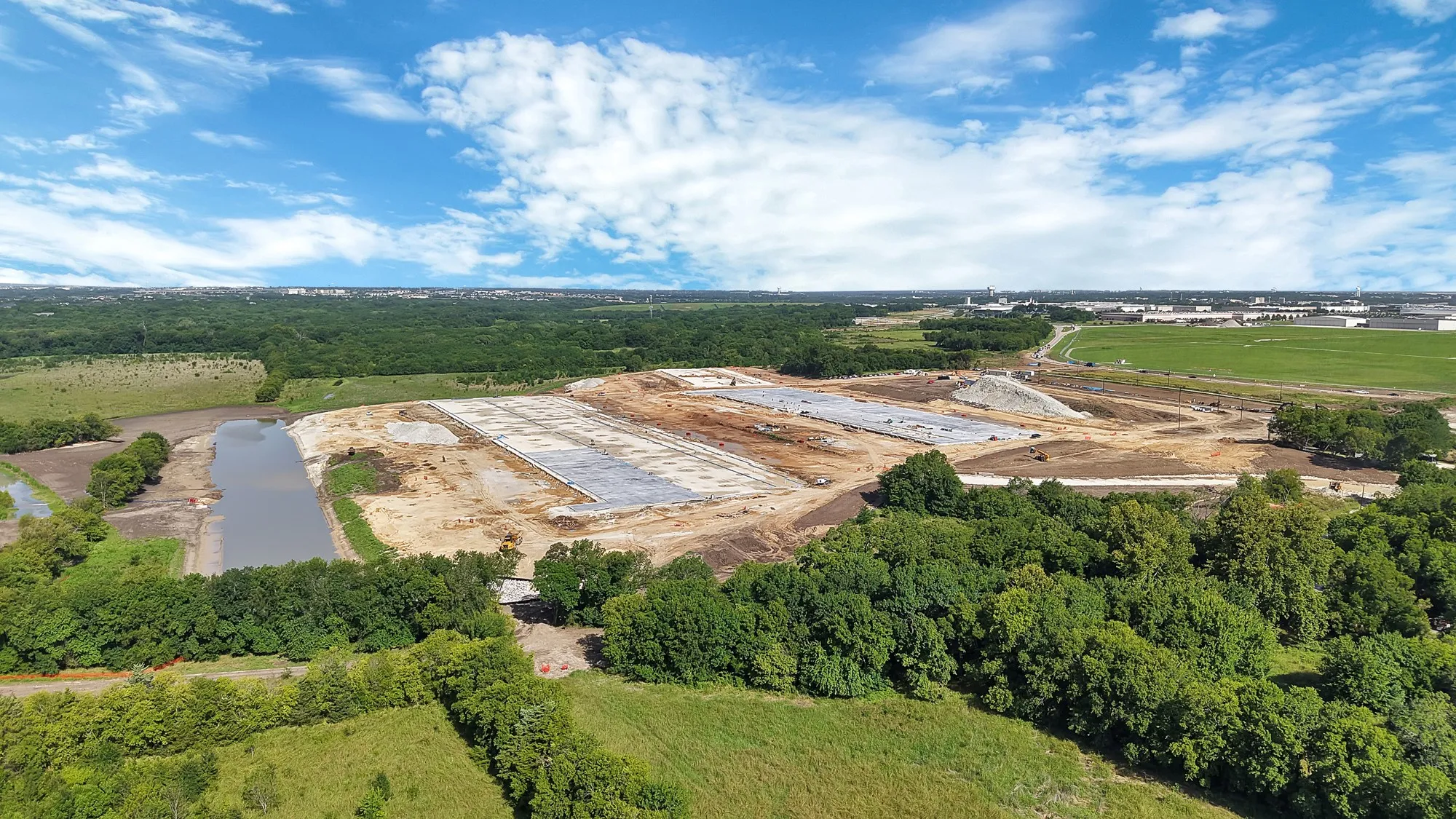 Expansive land parcel featuring multiple concrete foundation pads and ongoing site development, bordered by a substantial body of water and mature tree lines