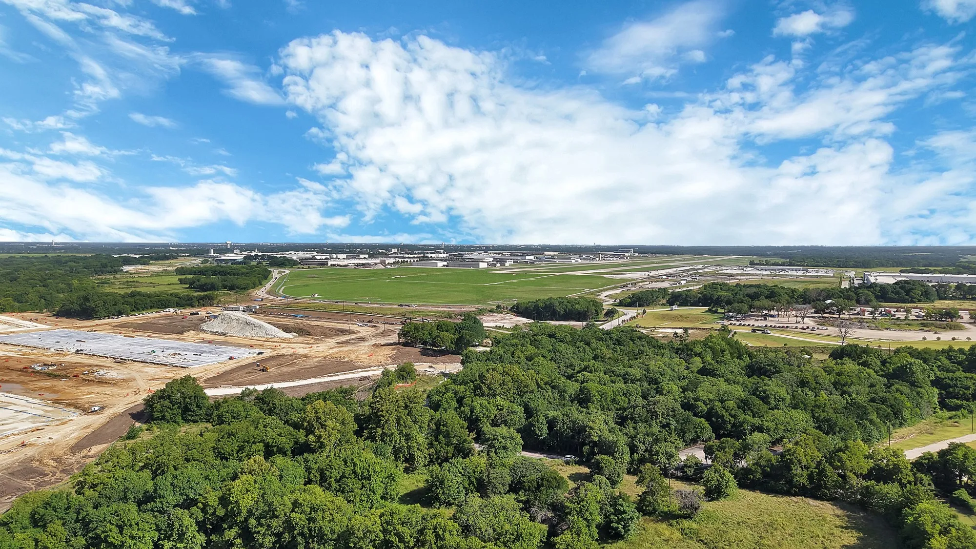 Expansive aerial landscape featuring a large wooded area, open green fields, and a distant industrial complex