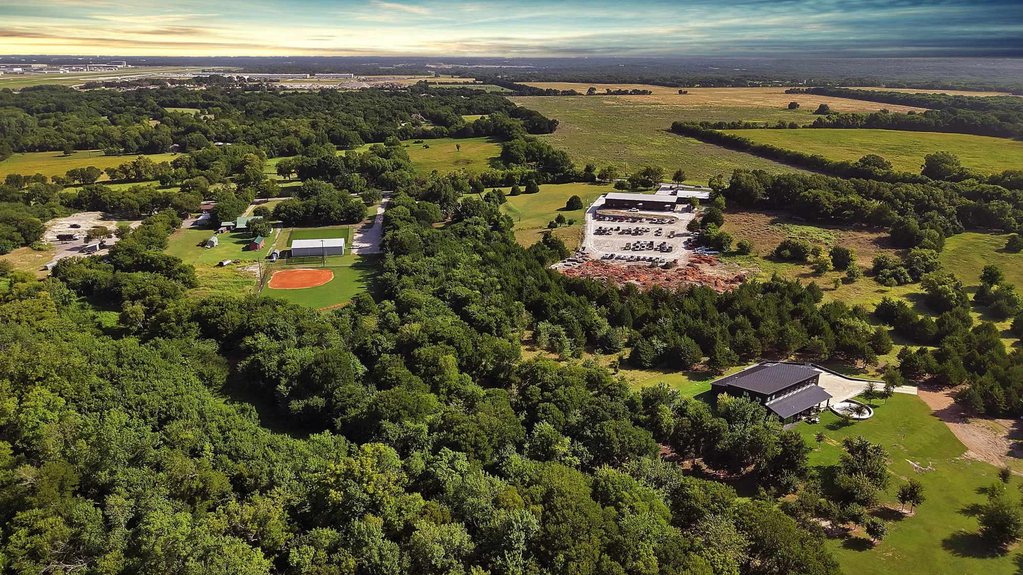 Expansive rural landscape featuring a large commercial building with extensive parking, a secondary building with a circular drive, a baseball diamond, and diverse tree coverage