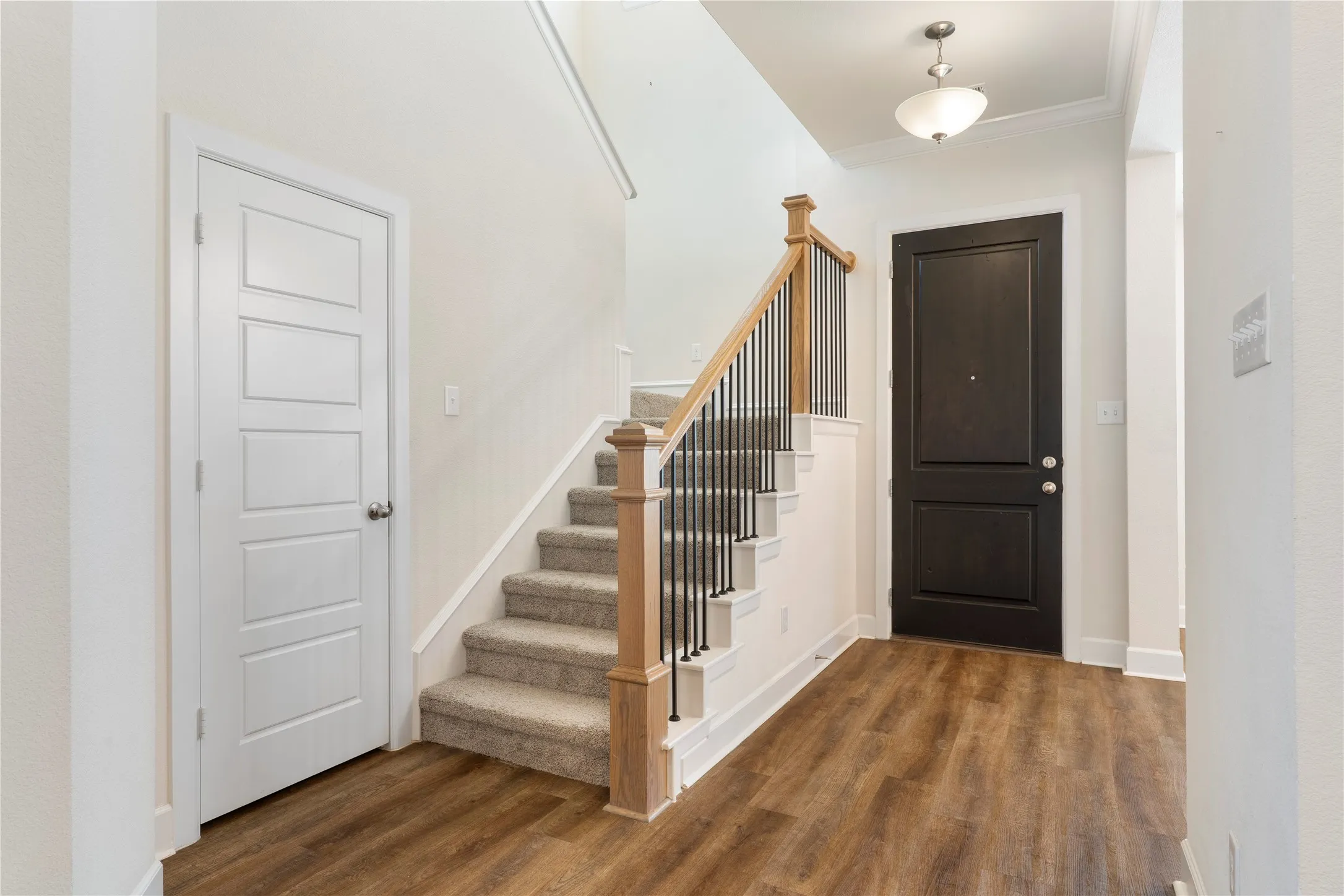 Wood-finish flooring, two-panel entry door, modern pendant light fixture, carpeted staircase with wood newel posts, and metal balusters
