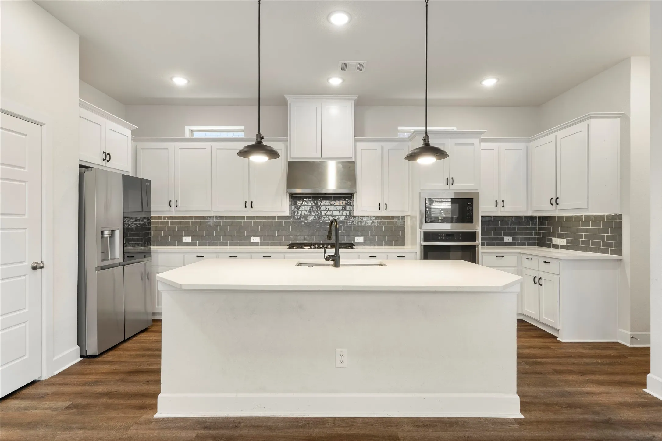 Central island with a light-toned countertop, white cabinetry with dark hardware, stainless steel appliances, grey subway tile backsplash, and wood-finish flooring
