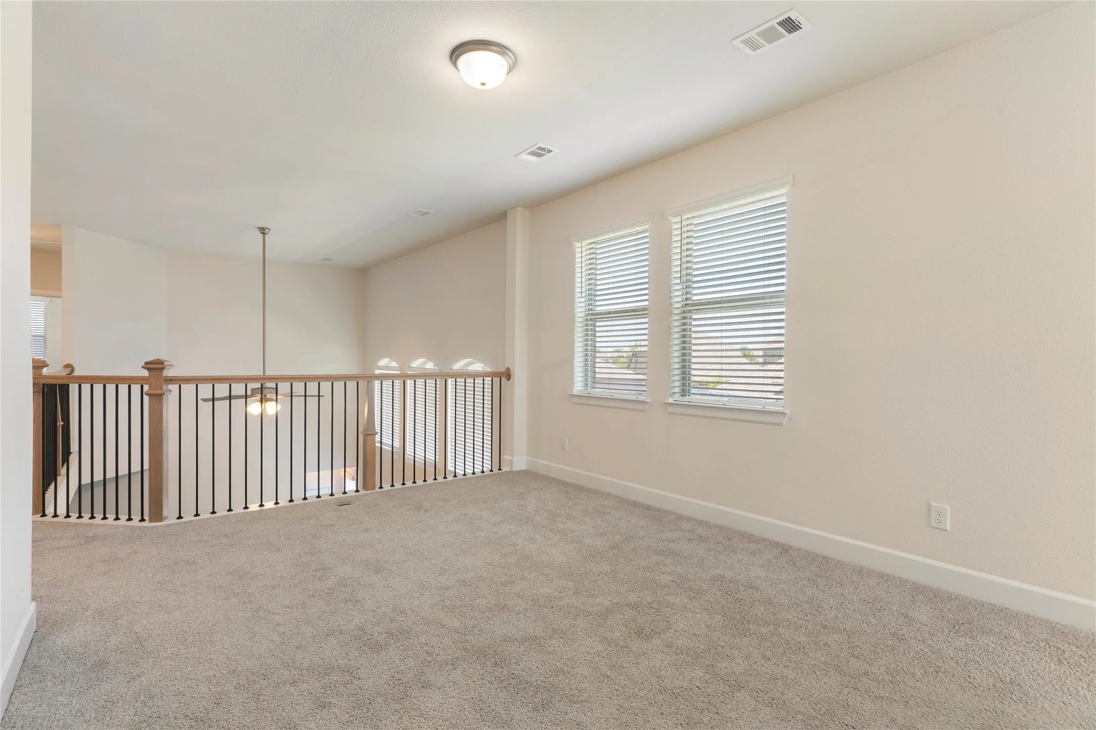 Carpeted loft area featuring two windows with blinds, a ceiling-mounted light fixture, and a wood and iron railing overlooking a lower space