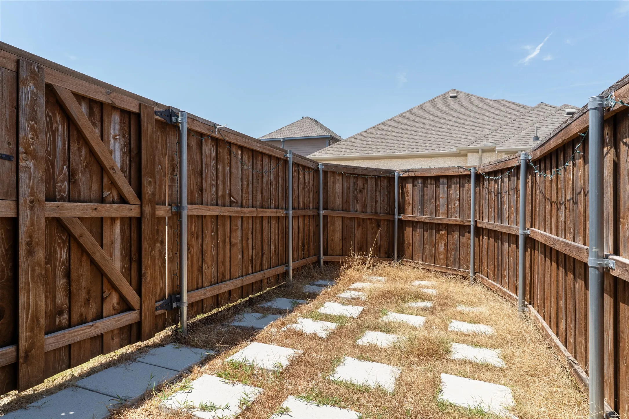Wood-stained privacy fencing with a gate, concrete stepping stones, and a shingle roofline visible