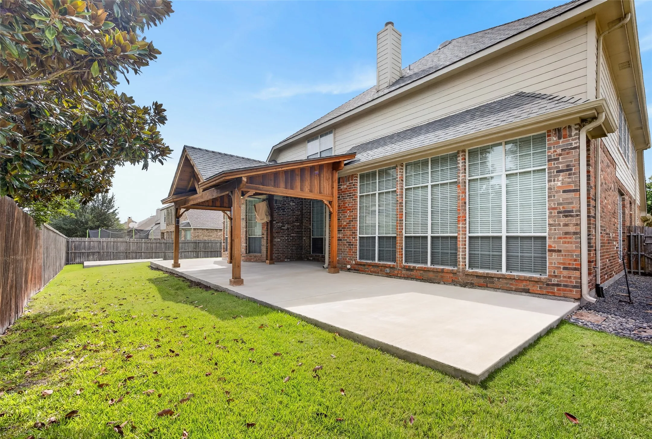 Expansive concrete patio featuring a wood-beamed pergola, brick exterior, multiple large windows, and a manicured lawn