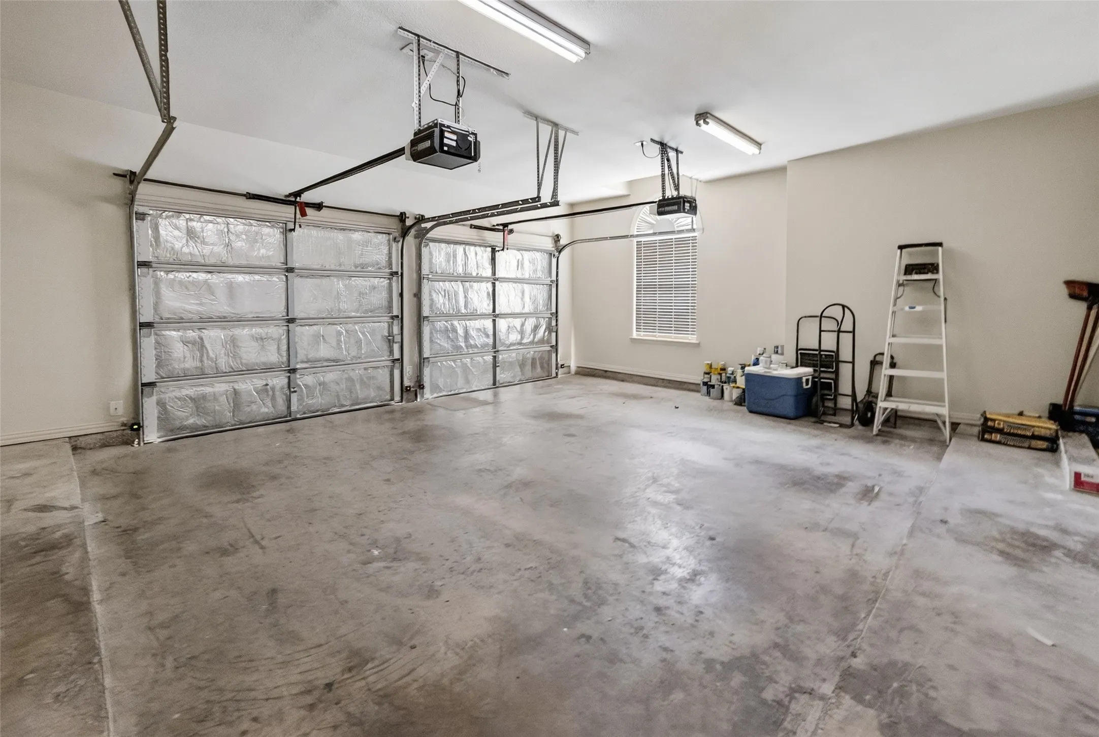 Spacious two-car garage featuring insulated overhead doors with automatic openers, overhead fluorescent lighting, a window with horizontal blinds, and painted walls
