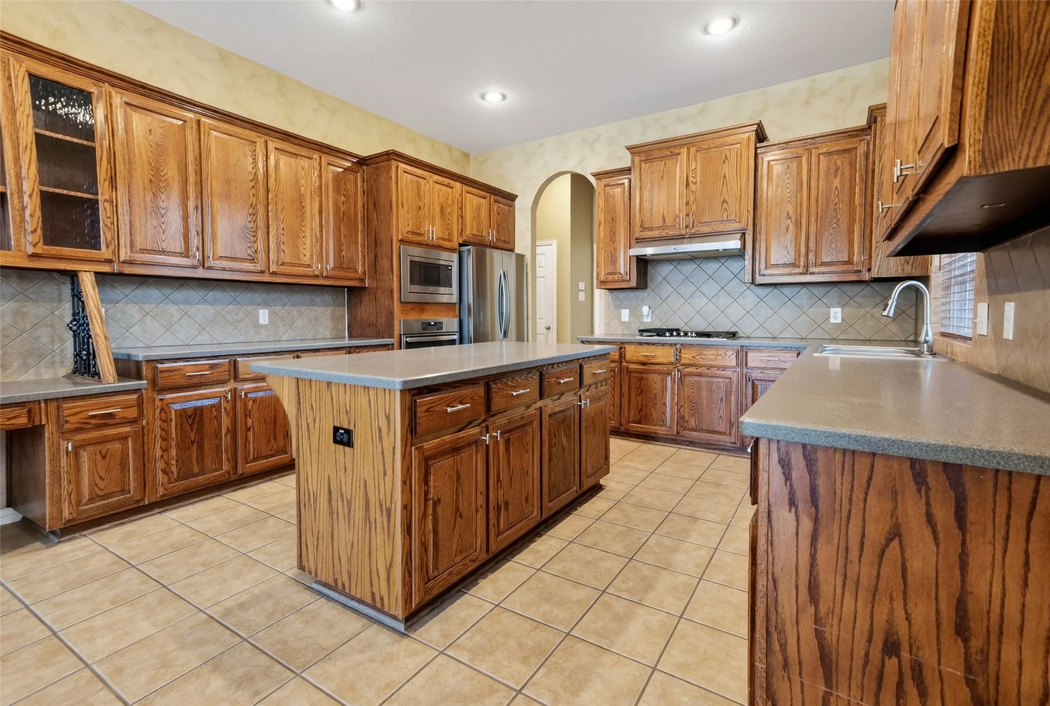 Spacious kitchen featuring extensive wood cabinetry, a central island with countertop, tile flooring, a tile backsplash, and recessed lighting