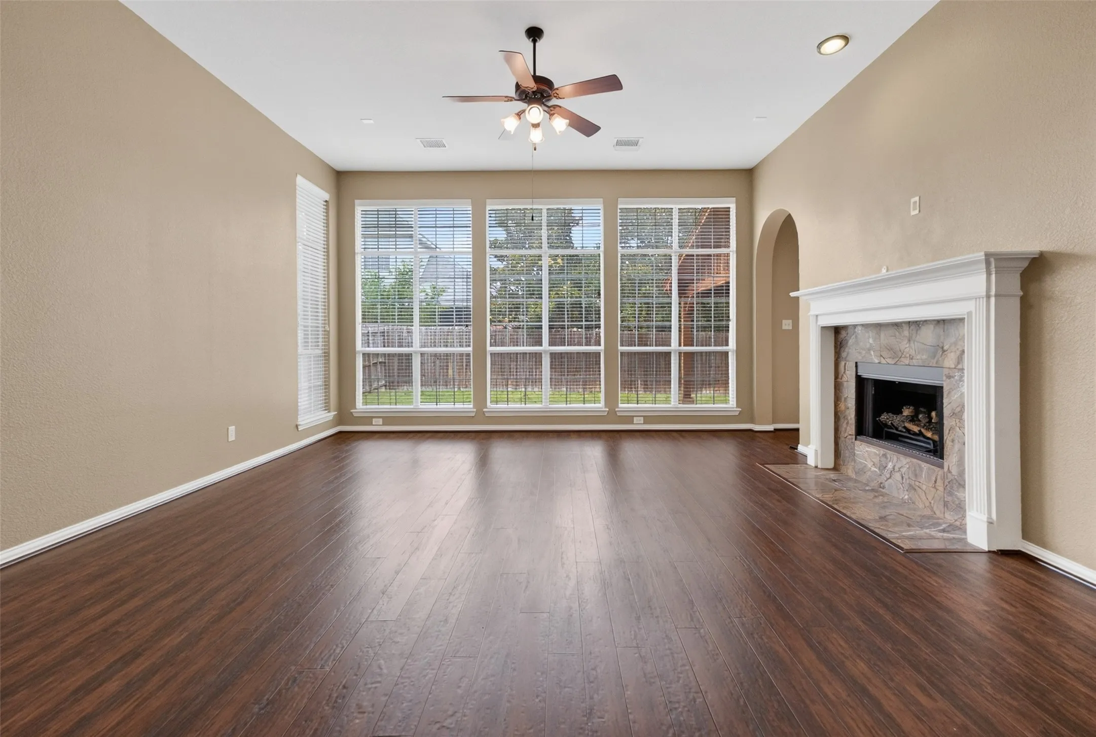 Spacious living area featuring wood-finish flooring, a decorative fireplace with a stone surround and white mantel, a ceiling fan with integrated lighting, and a wall of windows