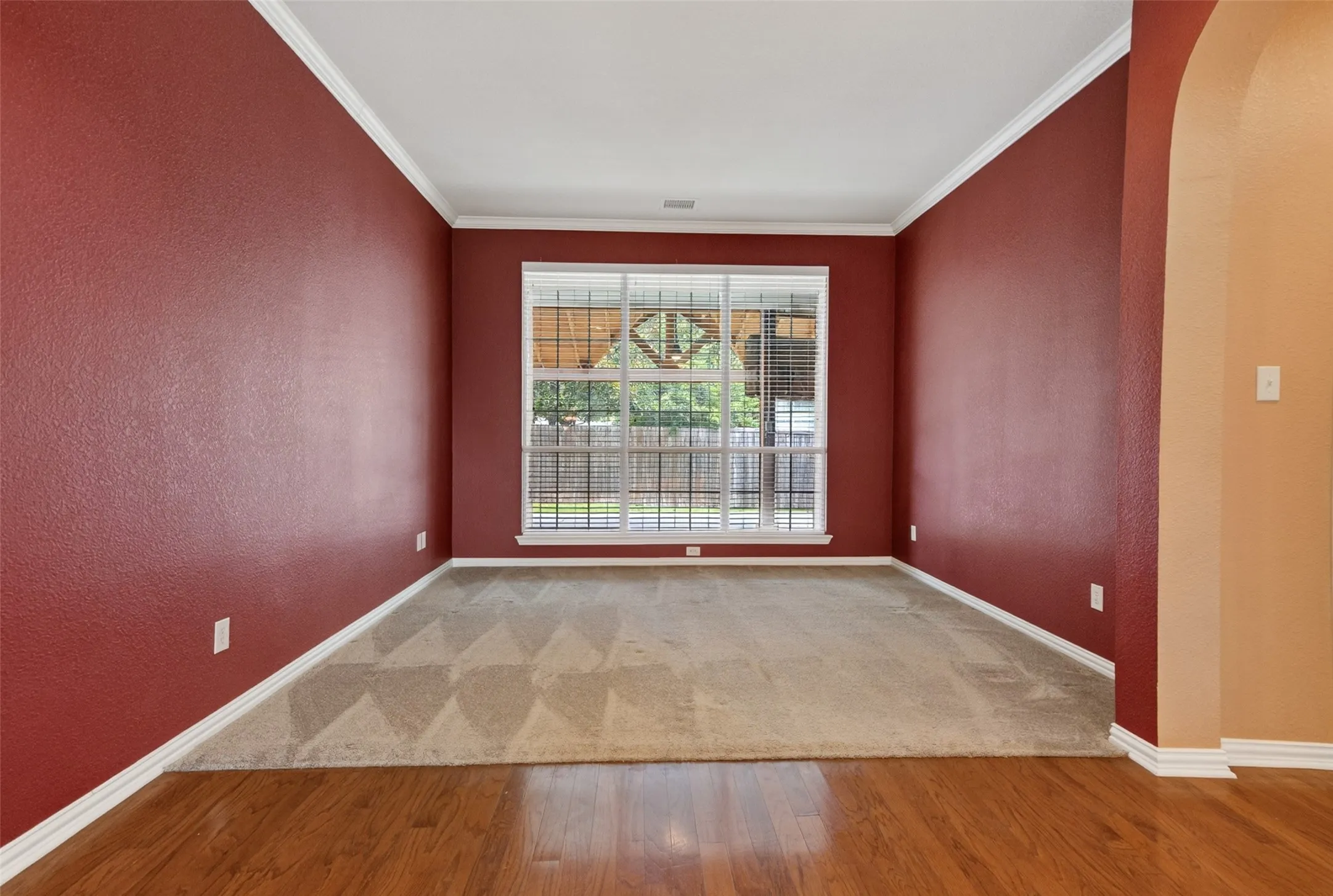 Hardwood flooring transitioning to carpeted area with a large window featuring horizontal blinds