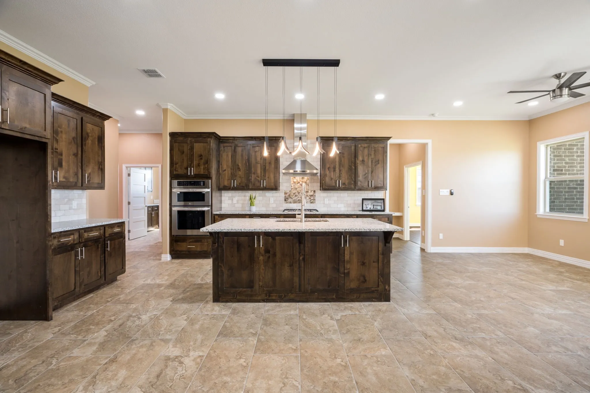 Spacious kitchen featuring an expansive center island with pendant lighting, rich wood-finish cabinetry, stainless steel double ovens, and a tile backsplash