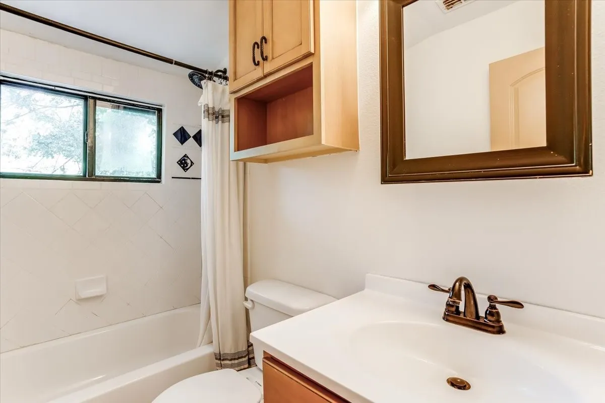 Bathroom featuring a white bathtub with tiled surround, a window, a single vanity with a bronze-finish faucet, and overhead cabinetry