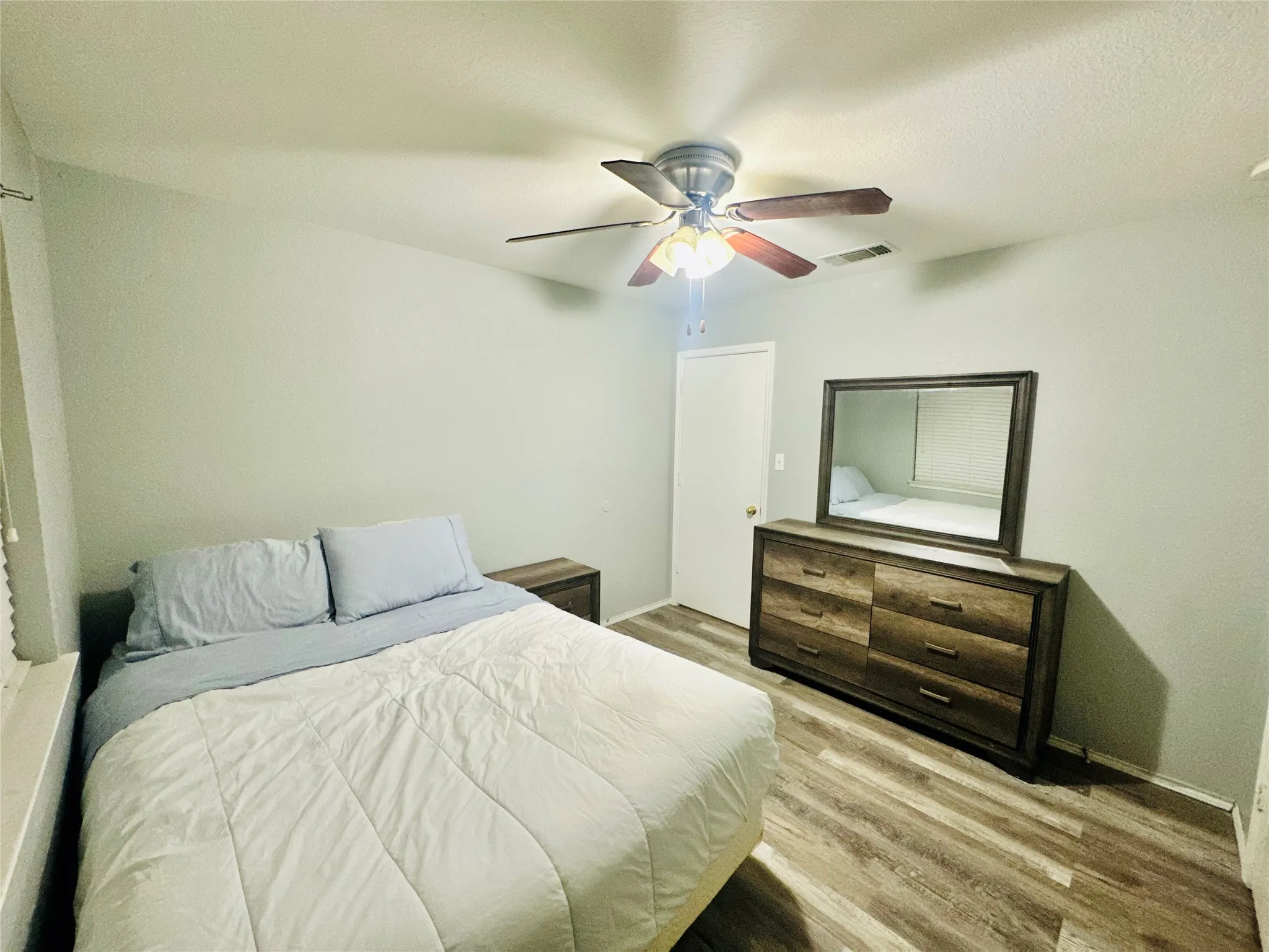 Wood-finish flooring with a neutral wall color, overhead ceiling fan with light fixture, and a white door with a brass-finish doorknob