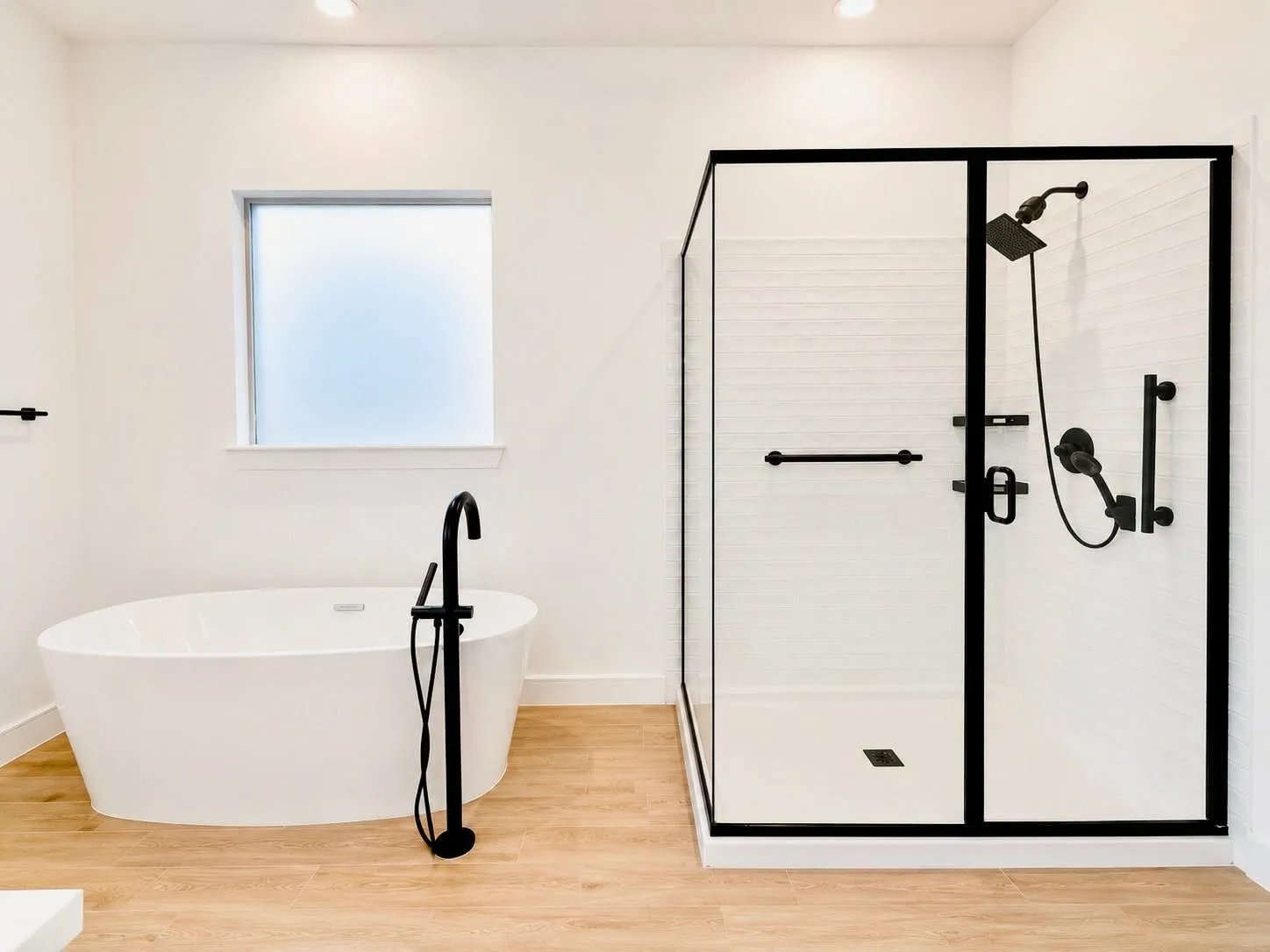 Modern bathroom featuring a freestanding soaking tub with a floor-mounted matte black faucet, a large walk-in shower with matte black framed glass, white subway tile shower surround, and light wood-finish flooring