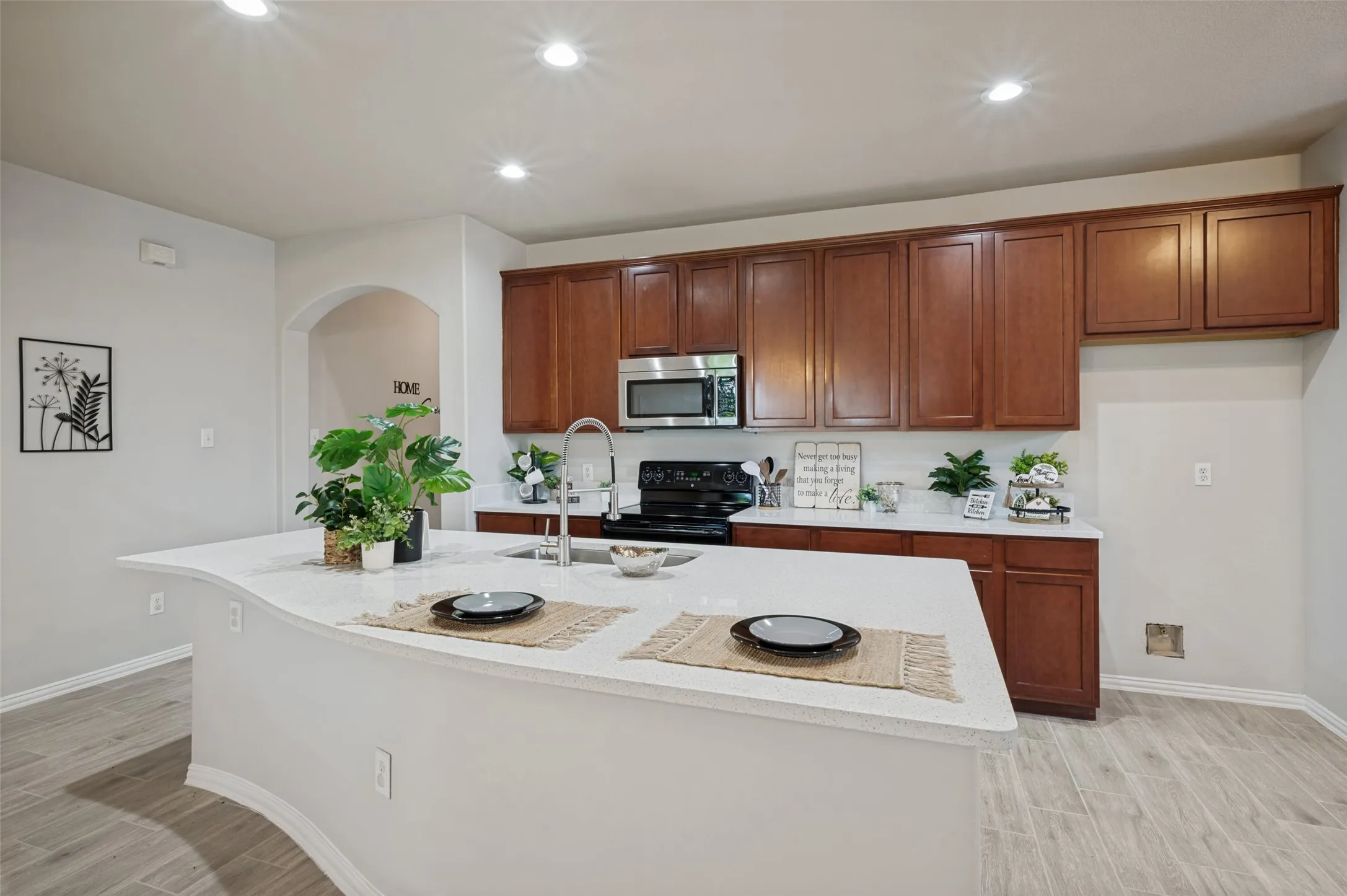 Kitchen featuring a large curved island with a double basin sink, light-toned countertops, rich wood-finish cabinetry, stainless steel microwave, and wood-finish flooring