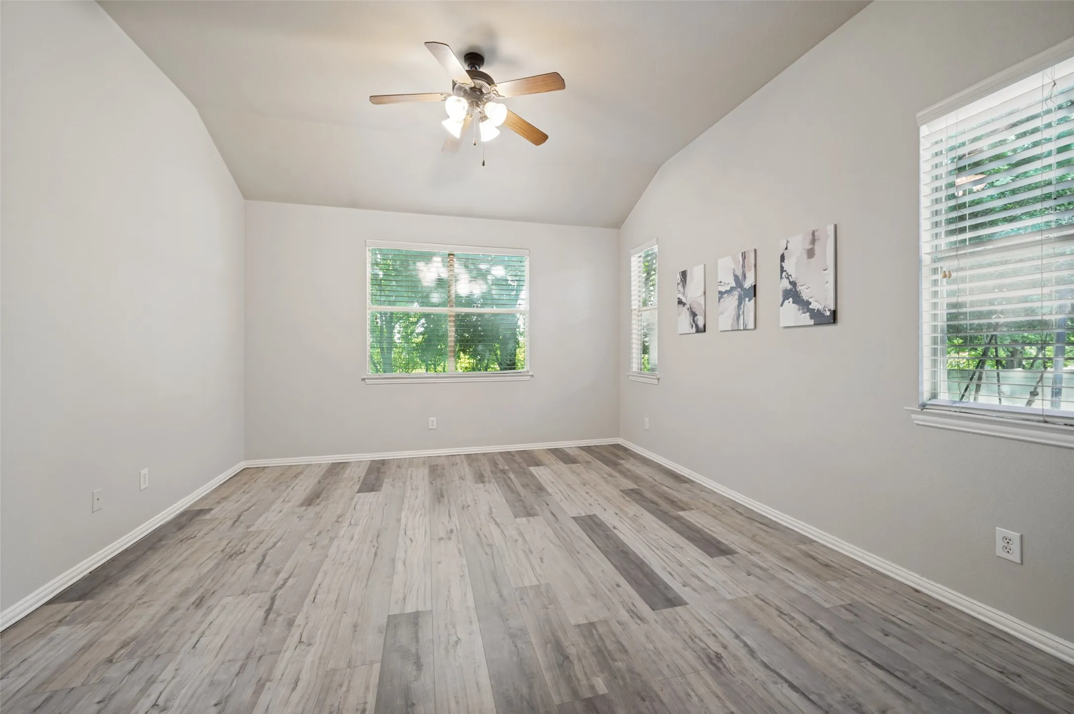 Vaulted ceiling room featuring wood-finish flooring, multiple windows with blinds, a ceiling fan, and light-toned walls
