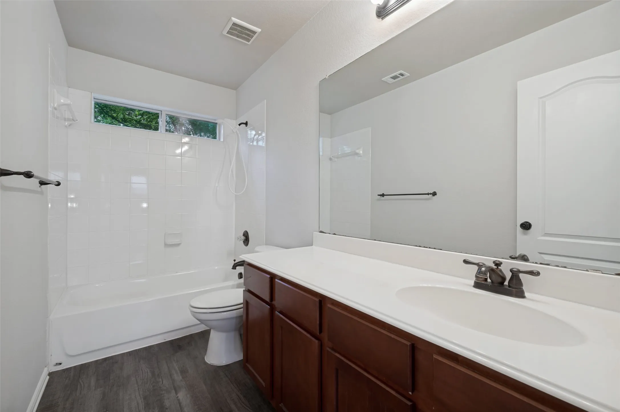 Bathroom featuring wood-finish flooring, a white vanity with a single basin sink, a large wall-mounted mirror, a white tiled tub/shower combination, and a horizontal window
