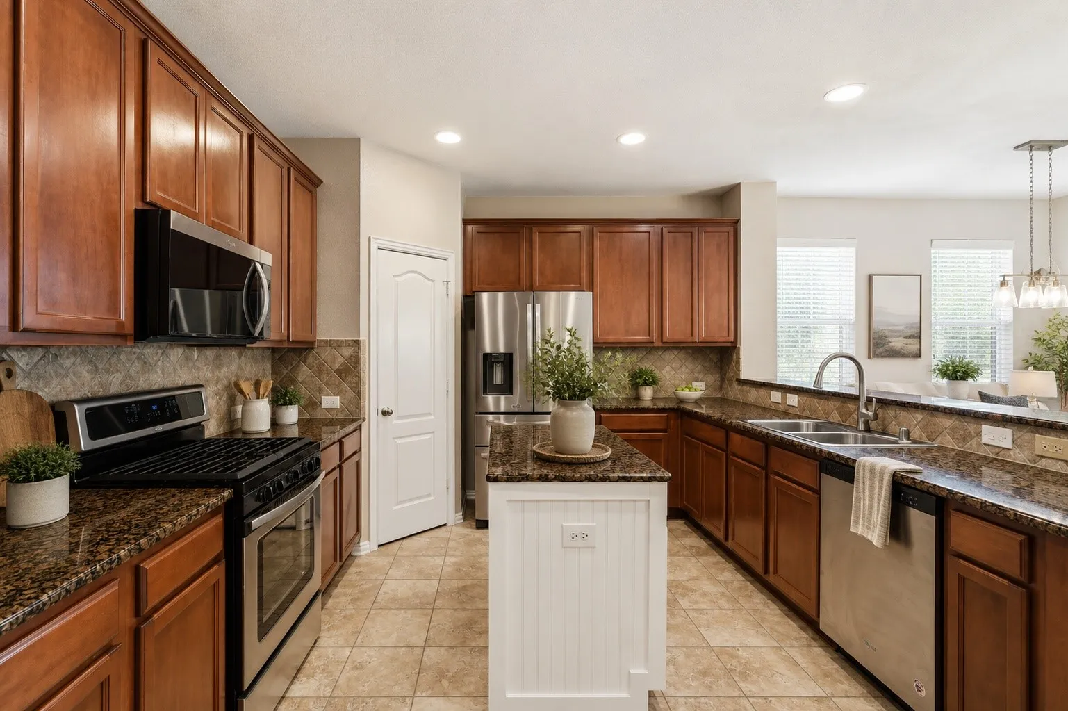 This spacious kitchen offers abundant cabinetry and generous granite countertop space, providing plenty of room for everyday meal prep and entertaining. Stainless steel appliances, a decorative tile backsplash, recessed lighting, and a center island add both function and style. A large double-basin sink is positioned beneath the windows for natural light, while the open design provides a seamless connection to the adjoining living and dining areas.