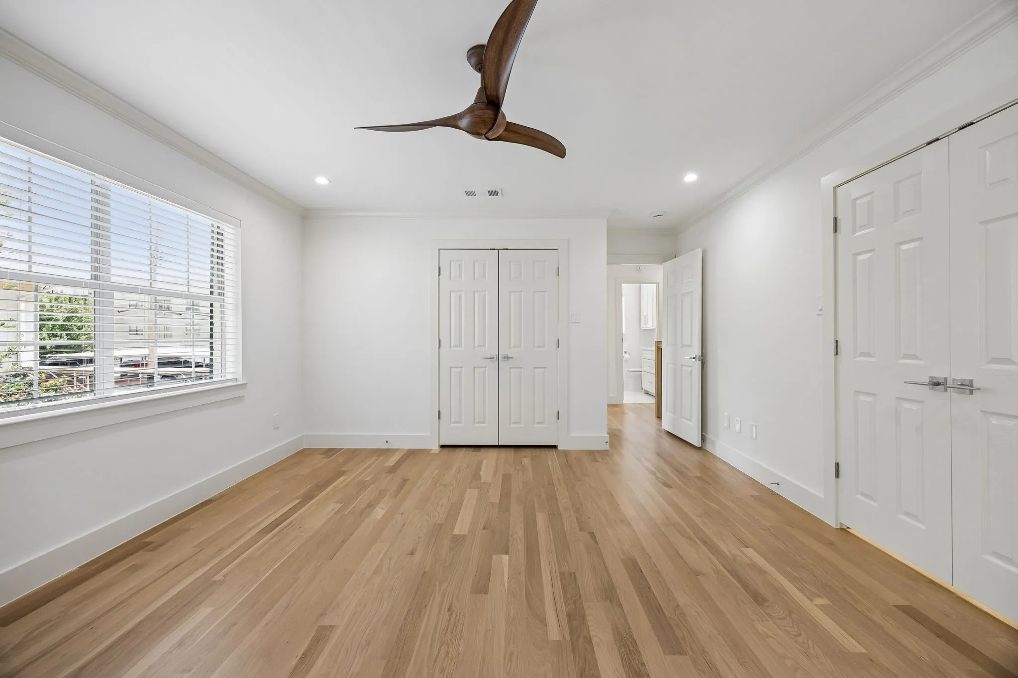Spacious room featuring light wood-finish flooring, a contemporary ceiling fan, and abundant natural light from a large window with blinds