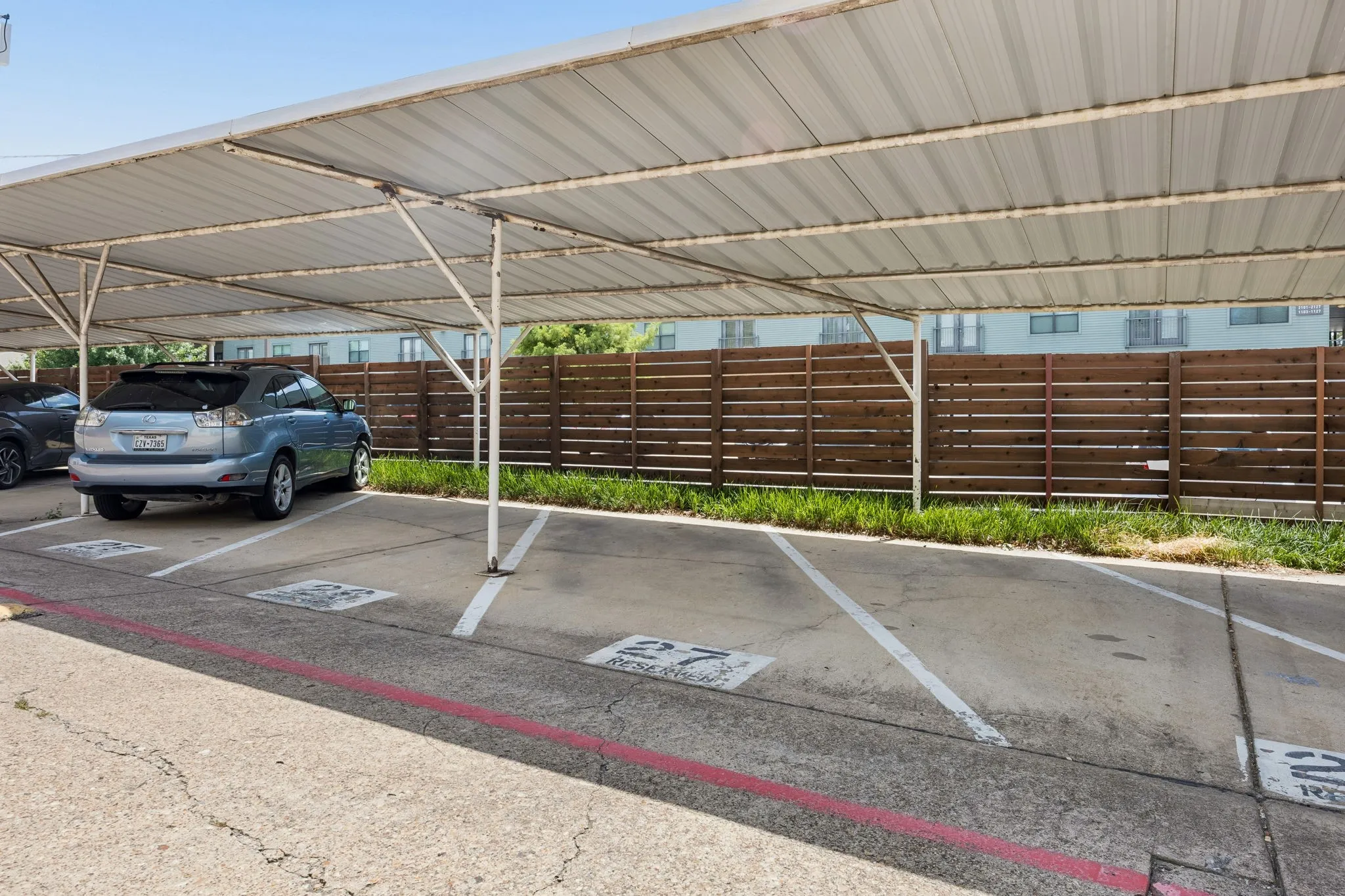 Covered parking structure with multiple spaces, featuring a metal roof and support pillars, concrete pavement, and a horizontal slat privacy fence