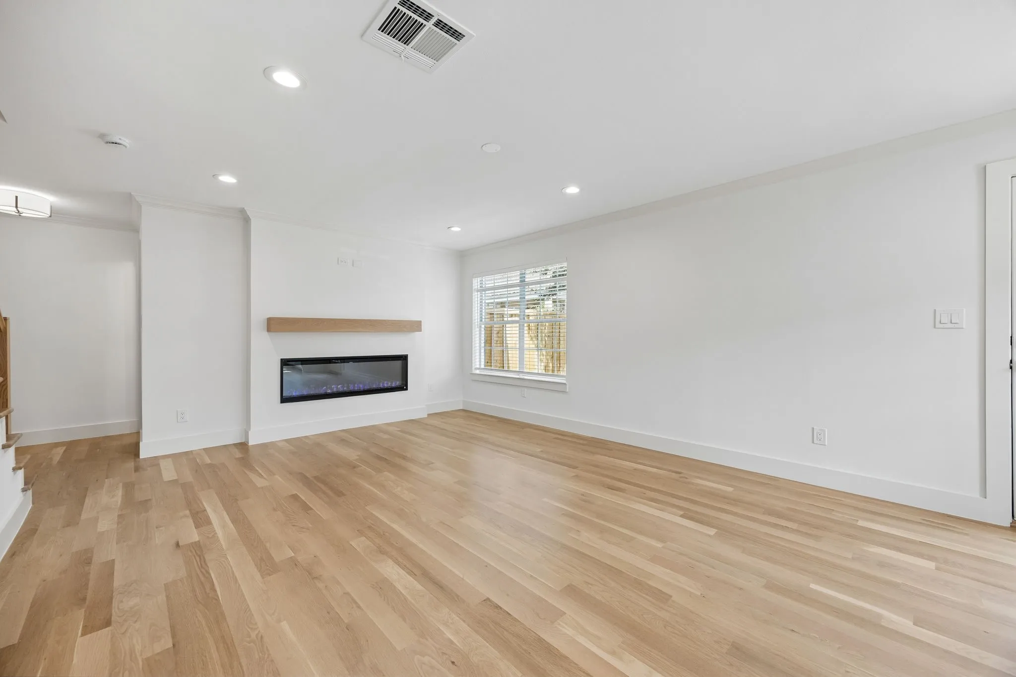 Spacious living area featuring light wood-finish flooring, a modern electric fireplace with a wood mantel, recessed lighting, and a large window providing natural illumination