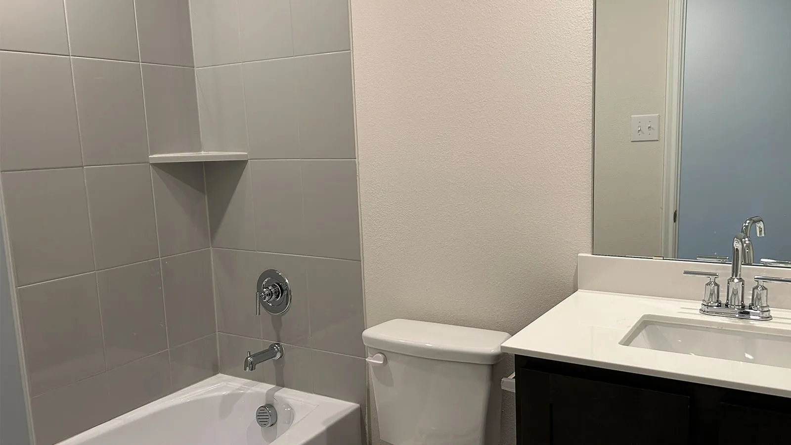 Full bathtub with a tiled surround featuring a built-in corner shelf, chrome fixtures, a single vanity with a white countertop, and an integrated sink