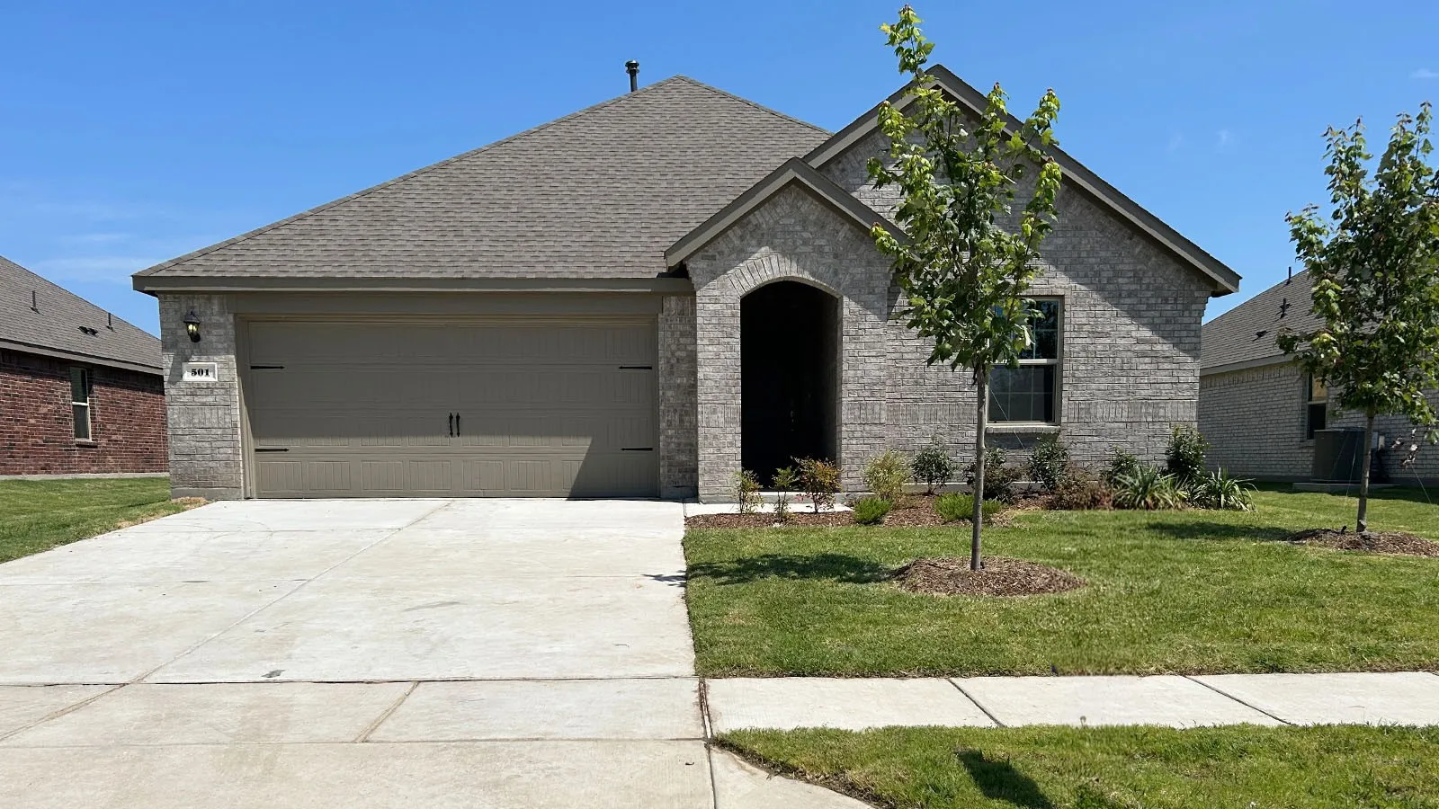 Brick facade with an arched entry, two-car garage, concrete driveway, and front lawn with young trees
