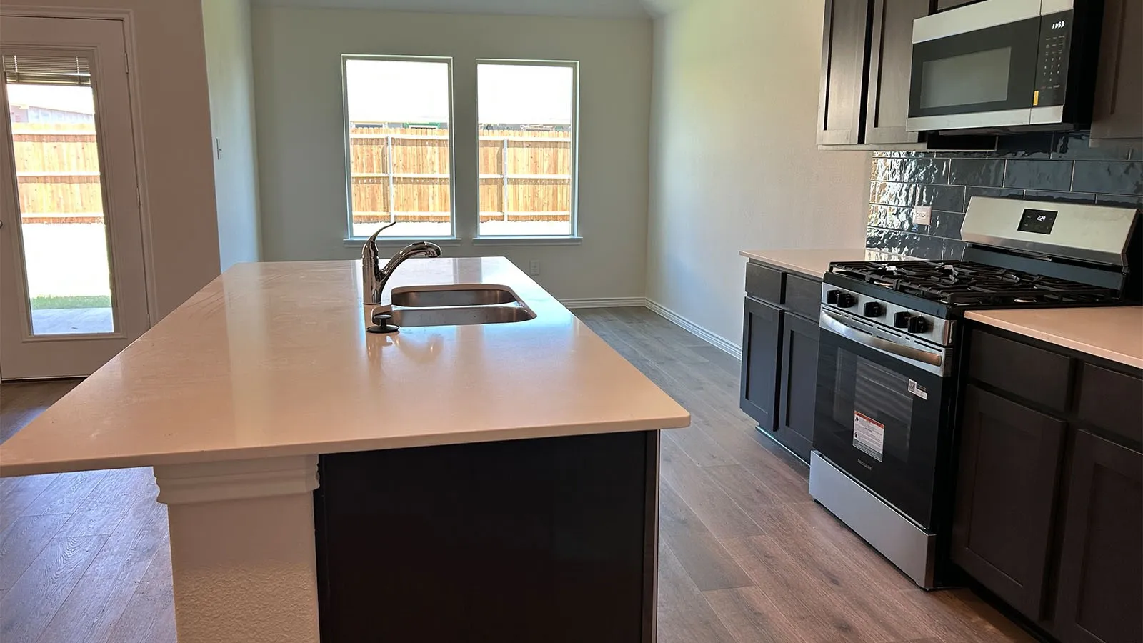 Kitchen featuring an island with a dual-basin sink, dark cabinetry, light countertops, wood-finish flooring, and stainless steel appliances