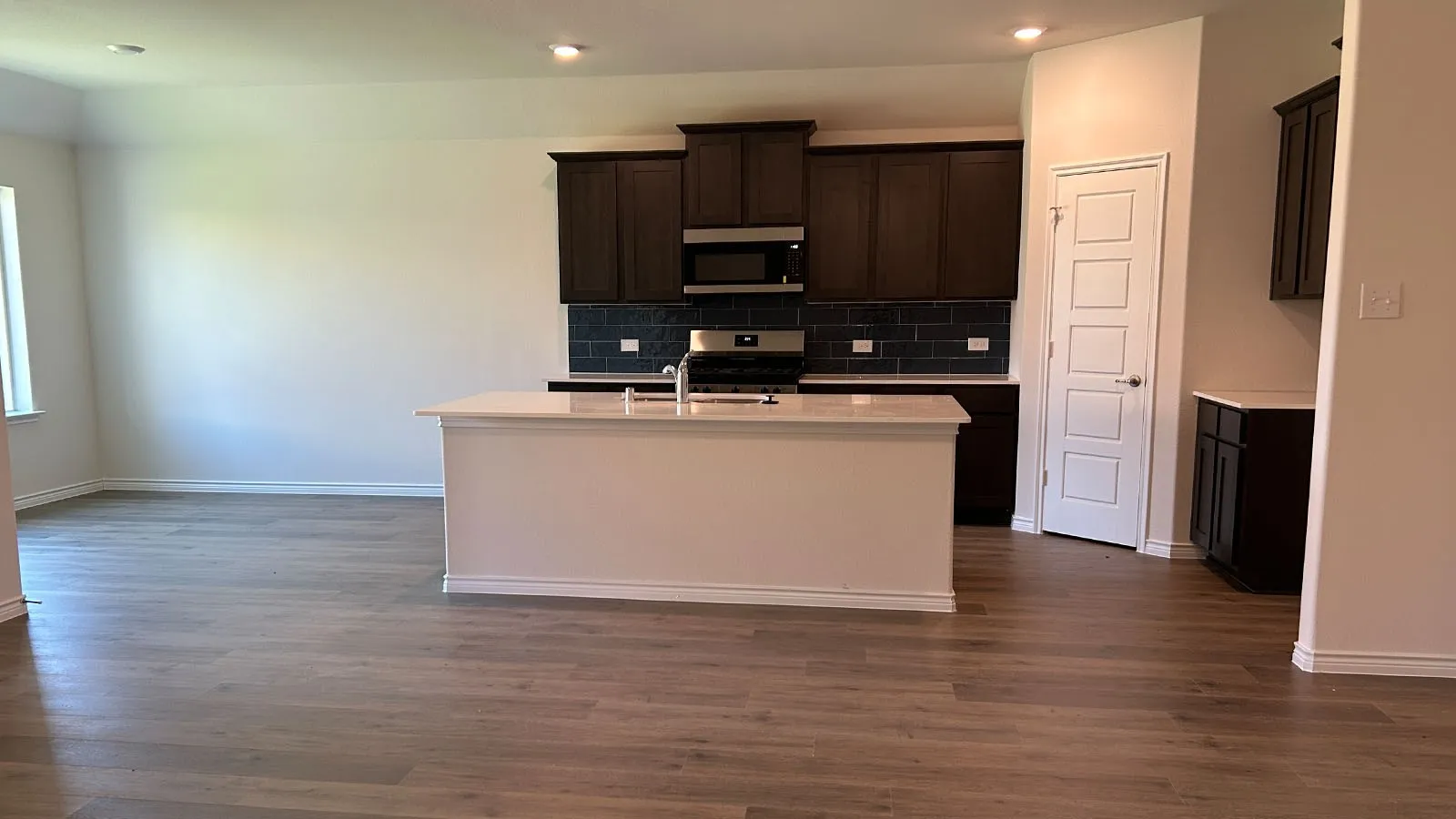 Expansive kitchen island with a light-toned countertop, dark wood-finish cabinetry, stainless steel appliances, dark subway tile backsplash, and wood-finish flooring