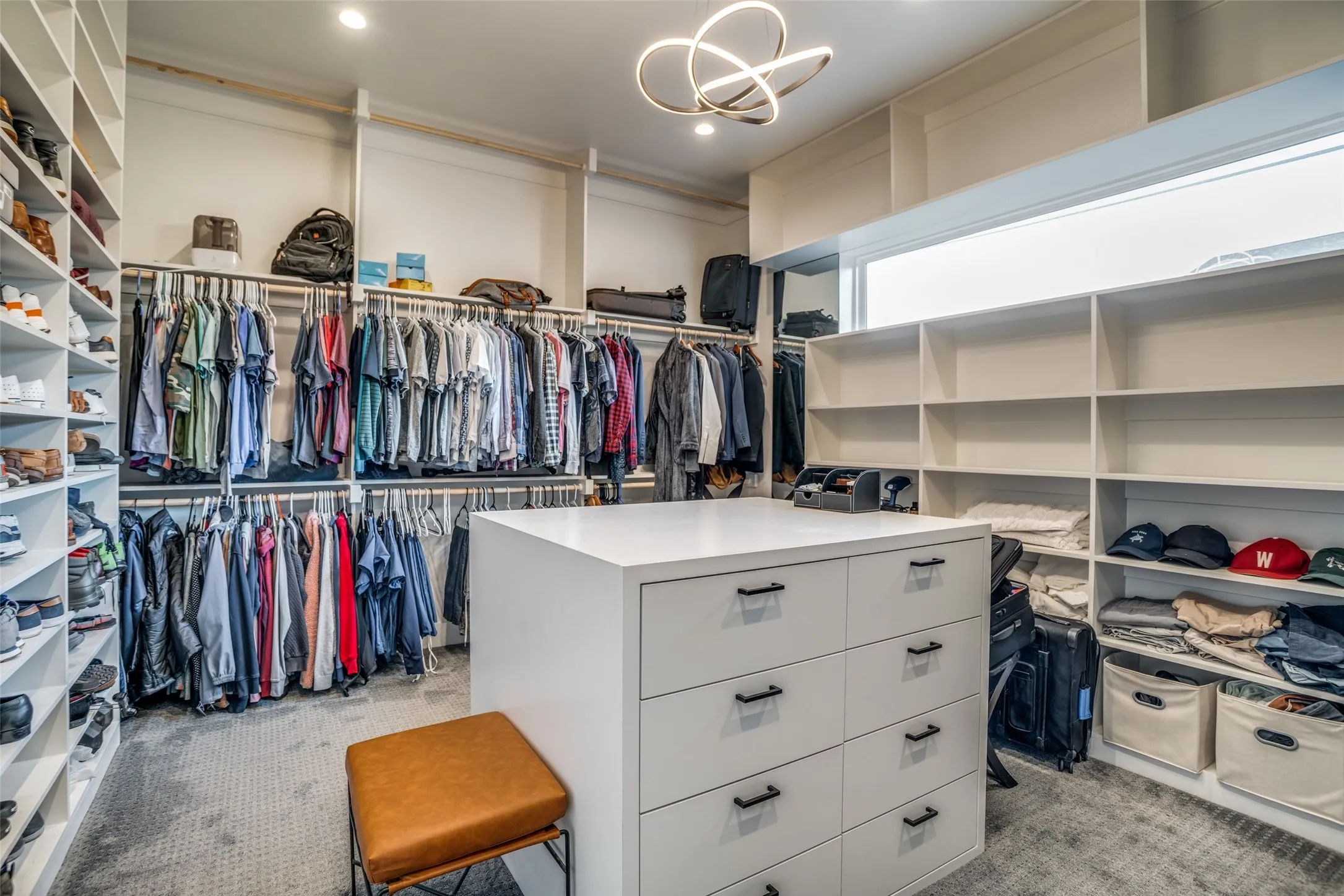 Spacious closet featuring a window providing natural light along with recessed lighting and a modern designer chandelier so you will never make the mistake of putting on 2 different colored shoes.  There is ample space for hanging clothes, shoes, bags and luggage and an island with extra drawer space.