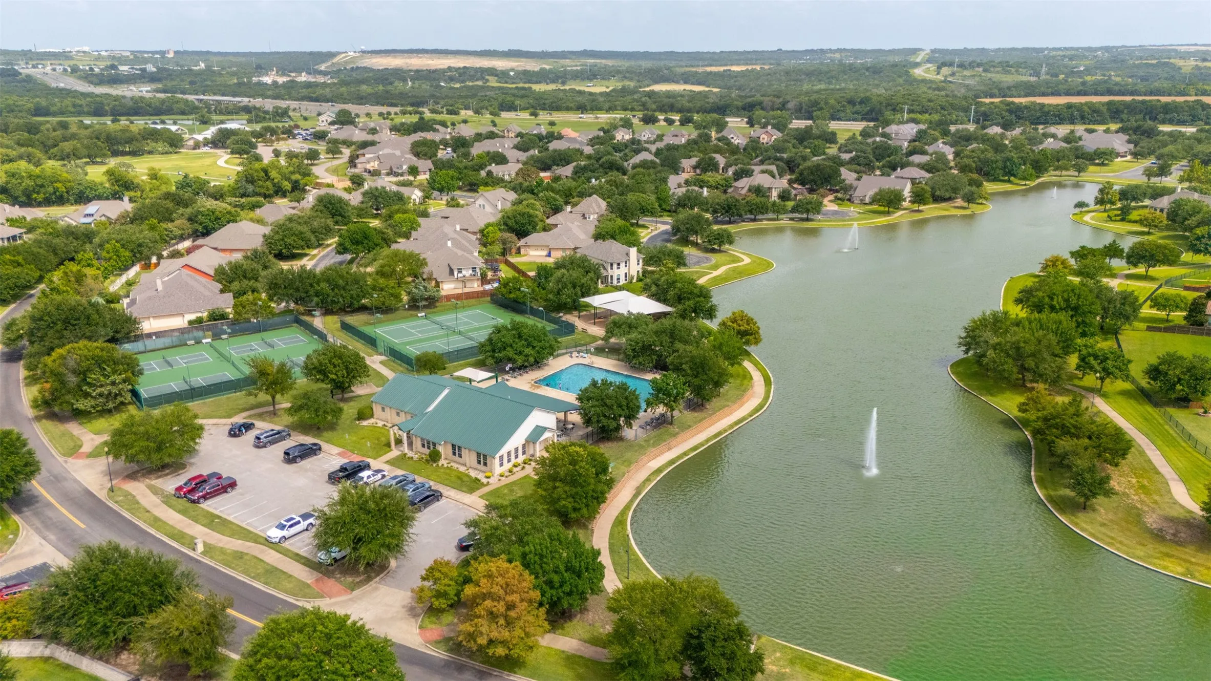 Aerial perspective showcasing a community with extensive green spaces, a large lake with fountains, multiple tennis courts, a swimming pool, and a community building