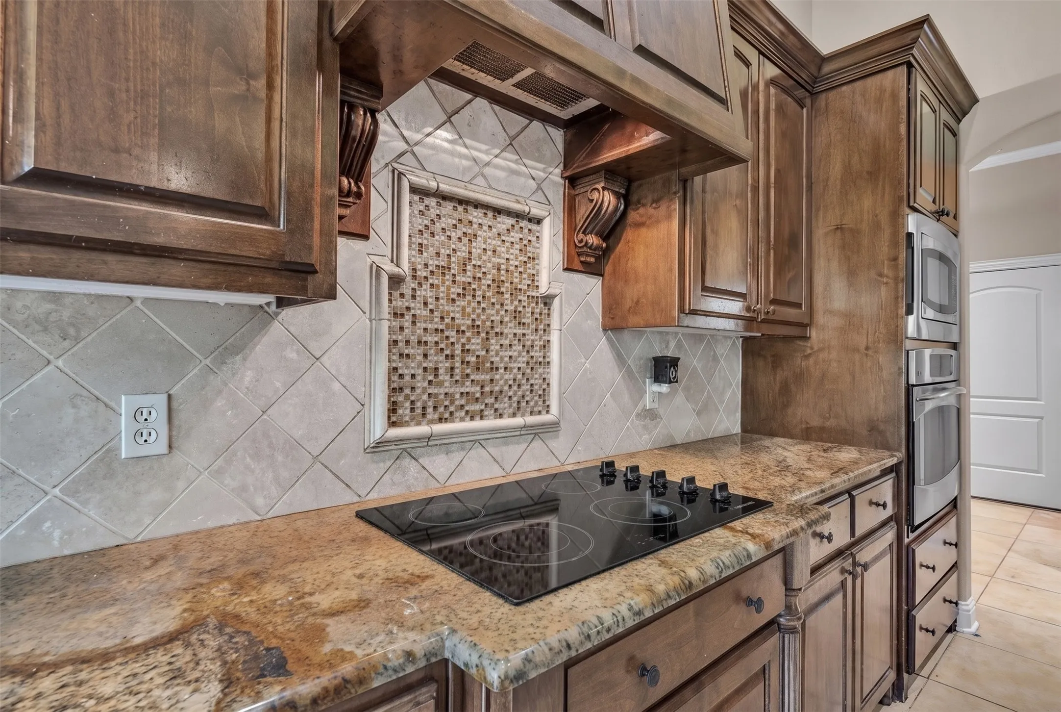 Kitchen featuring rich wood cabinetry, granite-finish countertops, a smooth cooktop, tile backsplash with decorative mosaic inlay, and built-in stainless steel appliances