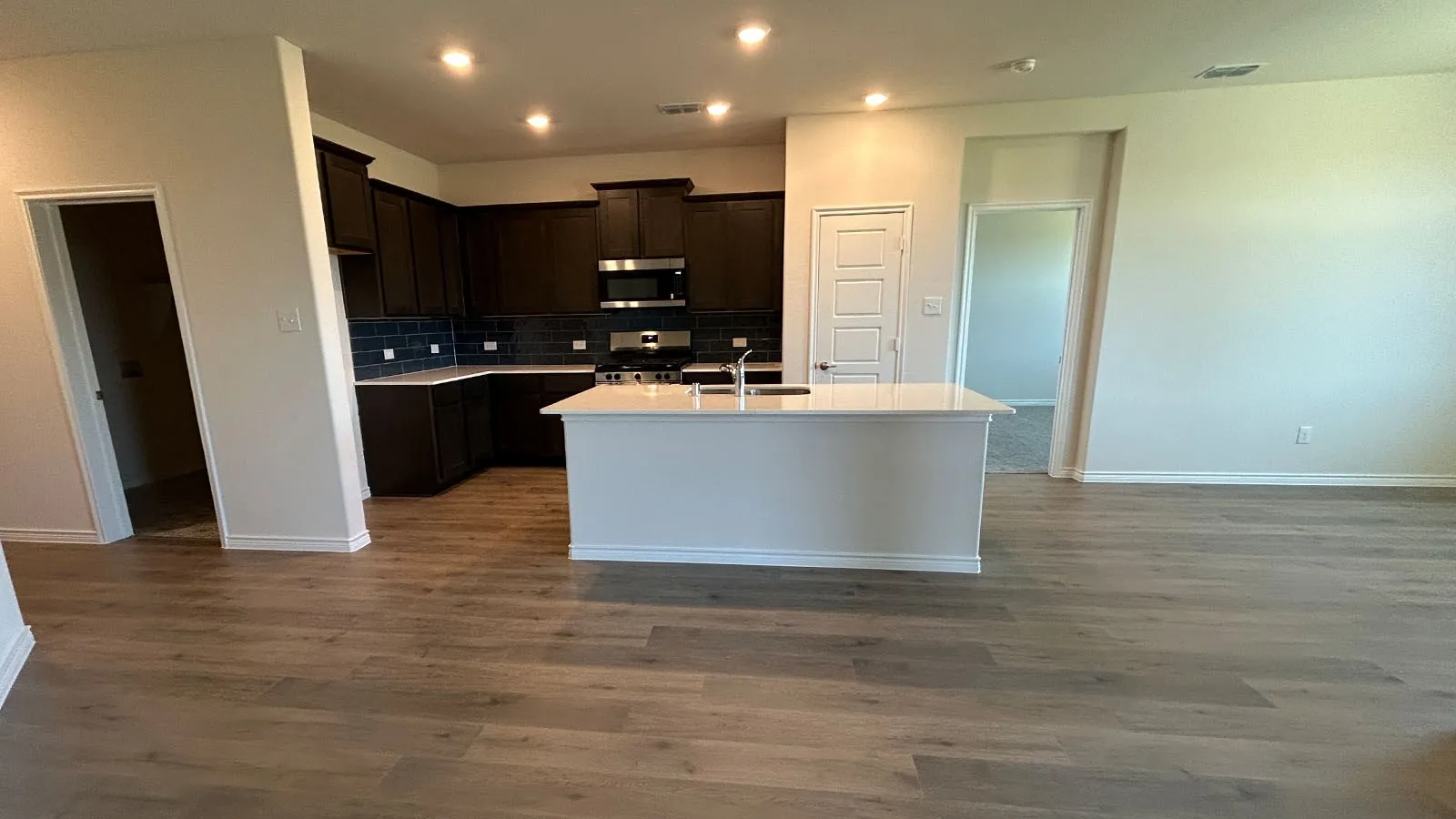 Wood-finish flooring throughout, kitchen featuring dark wood cabinetry, stainless steel appliances, light countertops, and a central island with an integrated sink