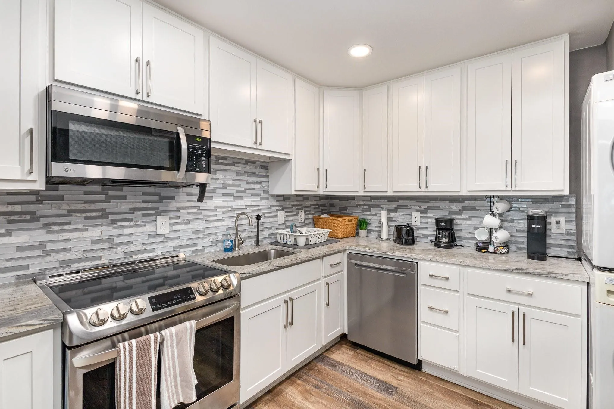 Modern kitchen featuring white shaker cabinetry, stainless steel appliances, a mosaic tile backsplash, stone-look countertops, and wood-finish flooring