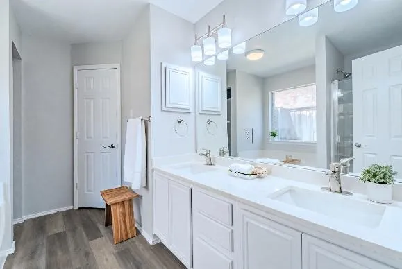 Dual vanity with integrated sinks and white cabinetry, large mirror with overhead lighting, wood-finish flooring, and white paneled doors