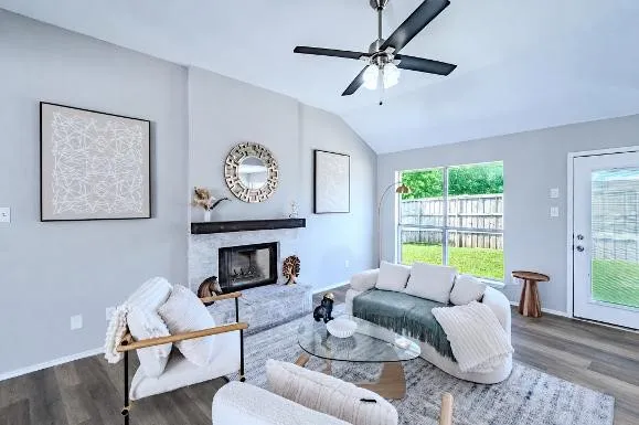 Vaulted ceiling with a ceiling fan, fireplace with a mantle, large window overlooking a fenced yard, and wood-finish flooring
