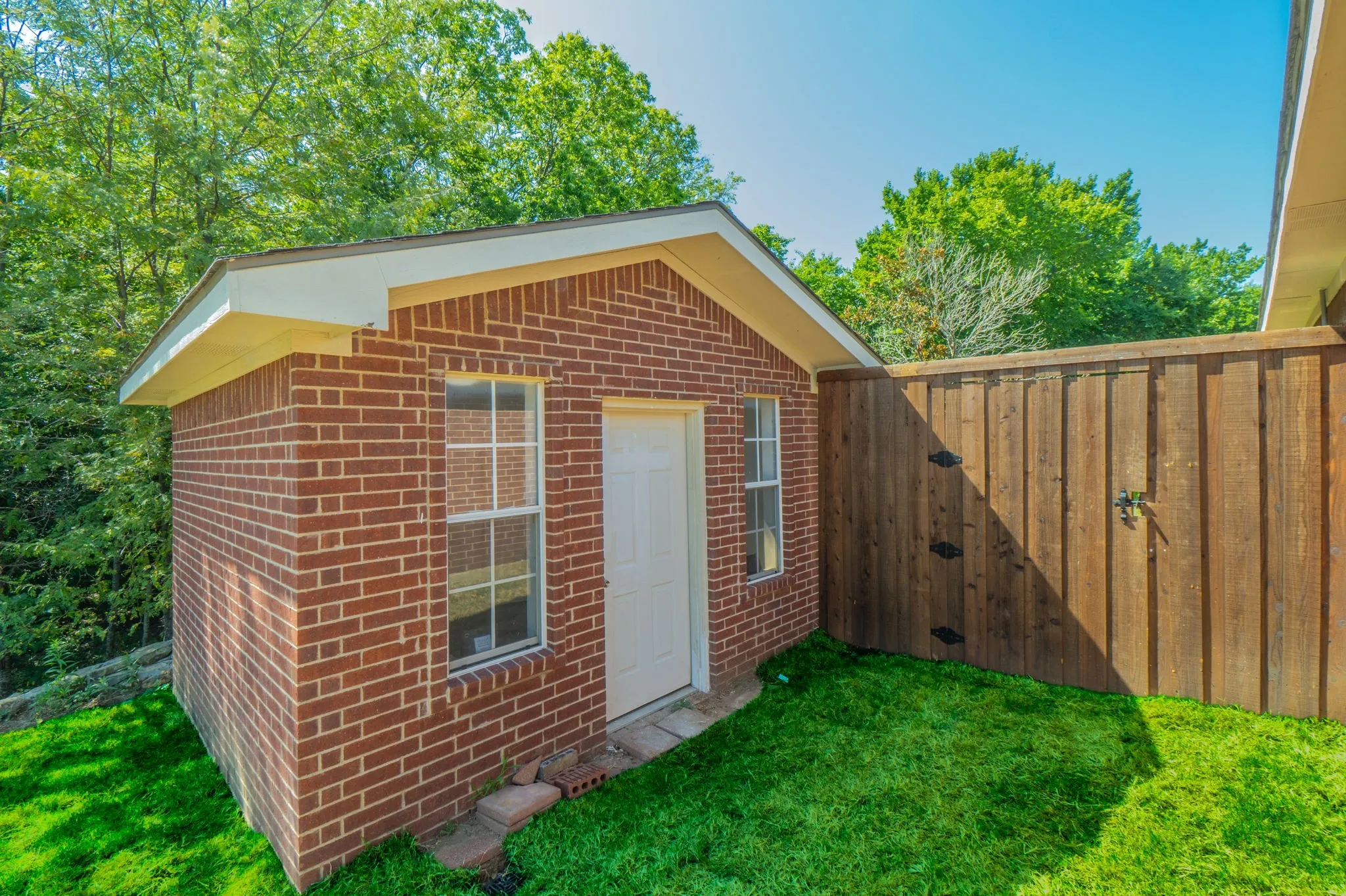 Brick outbuilding with a white door and two windows, featuring a sloped roofline and a surrounding grassy area