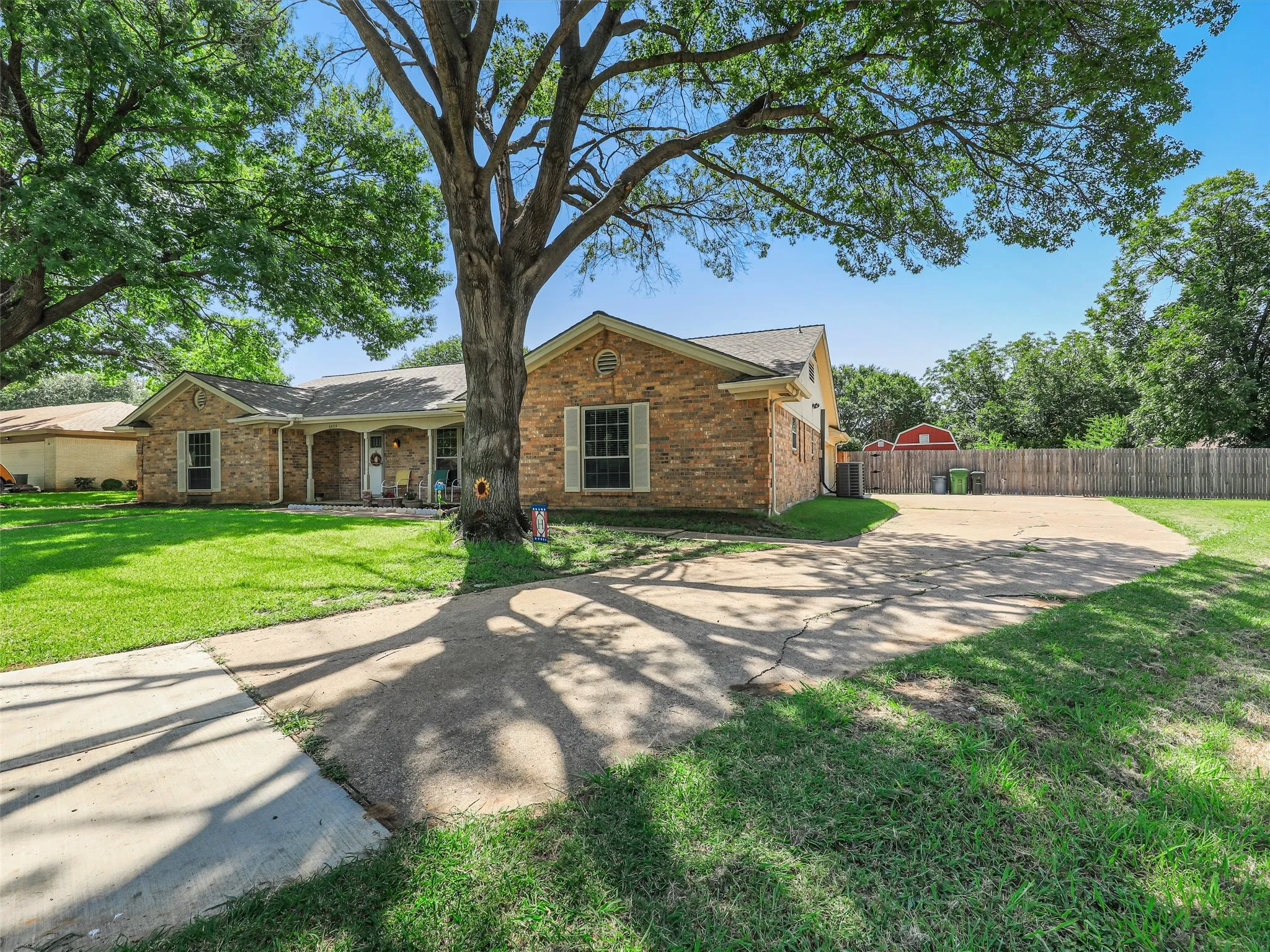 Brick facade home featuring a covered front porch, a concrete driveway, established trees, and a wooden privacy fence