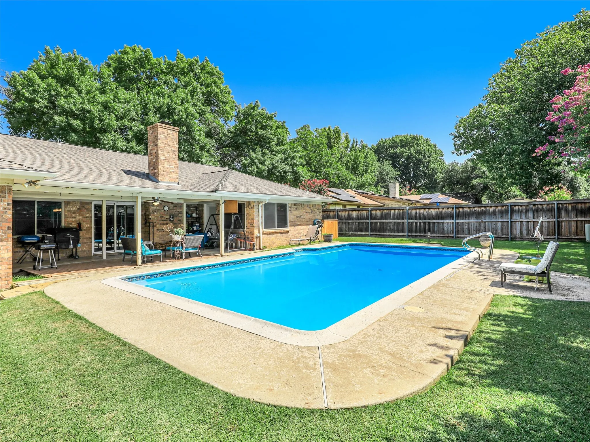 Backyard featuring a rectangular swimming pool with concrete surround, expansive lawn, and a covered patio with brick exterior