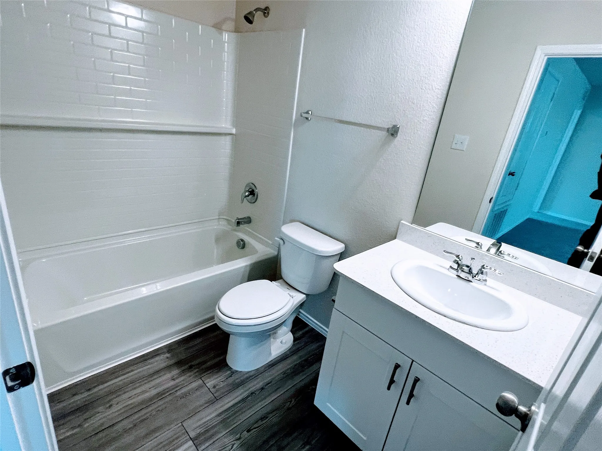 Bathroom featuring white subway tile shower surround, chrome fixtures, dark wood-finish flooring, white vanity with integrated sink, and a speckled white countertop
