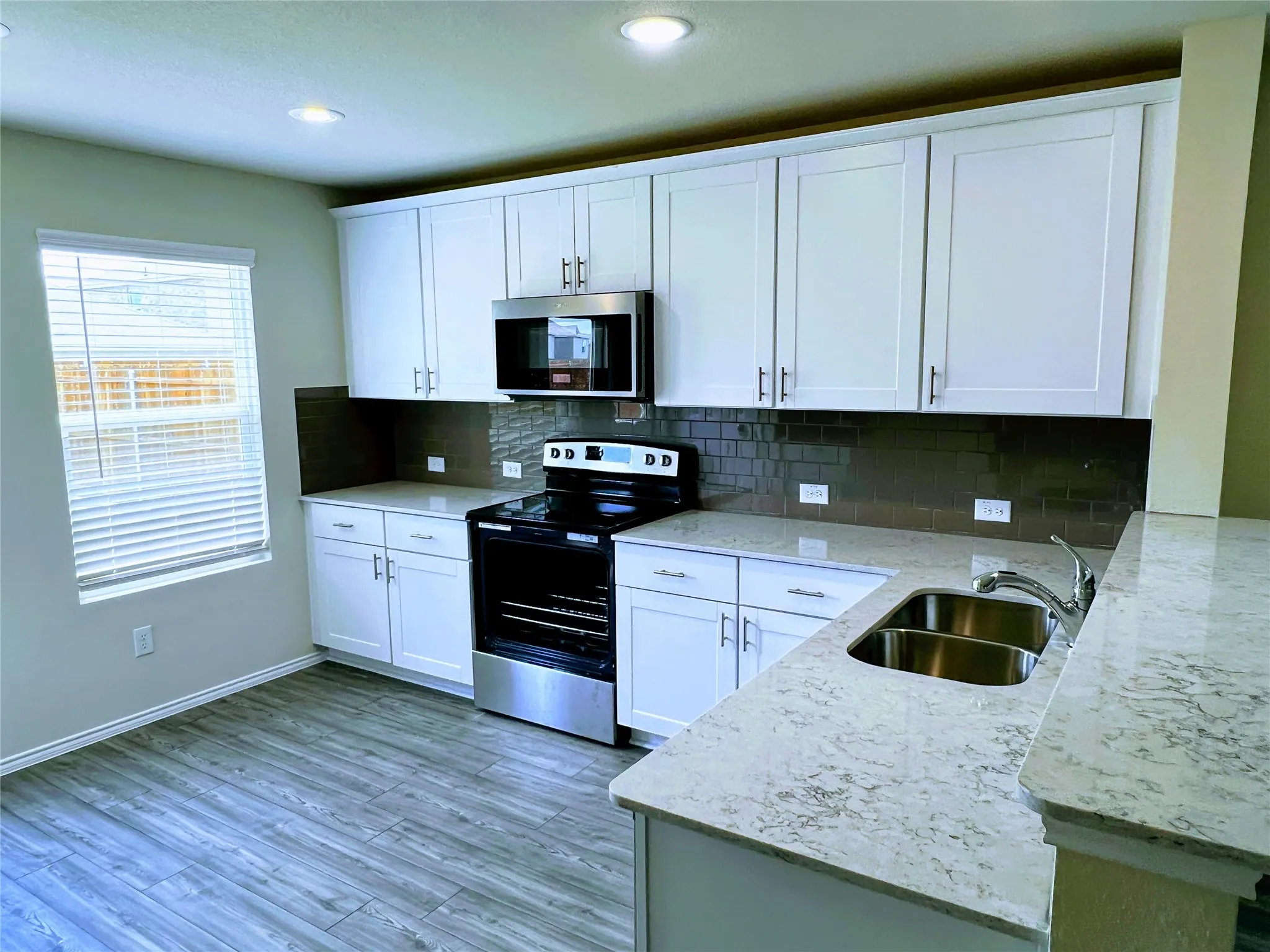 Kitchen featuring white shaker-style cabinetry, light-toned countertops, a stainless steel double-basin sink, and a dark subway tile backsplash