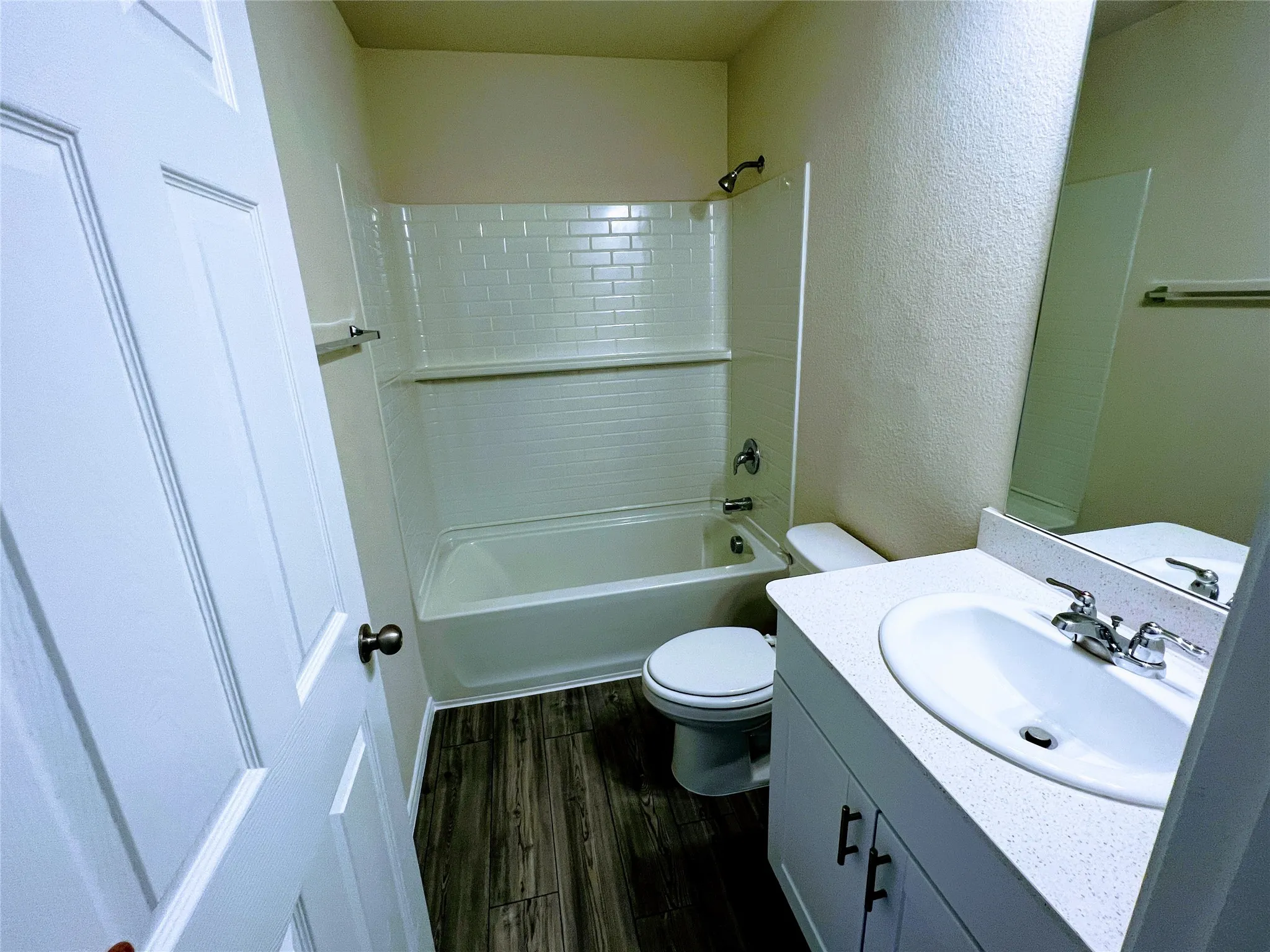 Bathroom featuring a white vanity with an integrated sink, dark wood-finish flooring, a white bathtub with subway tile surround, and a toilet
