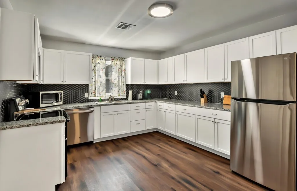 Kitchen featuring white shaker cabinetry, stainless steel appliances, dark hexagonal tile backsplash, granite-finish countertops