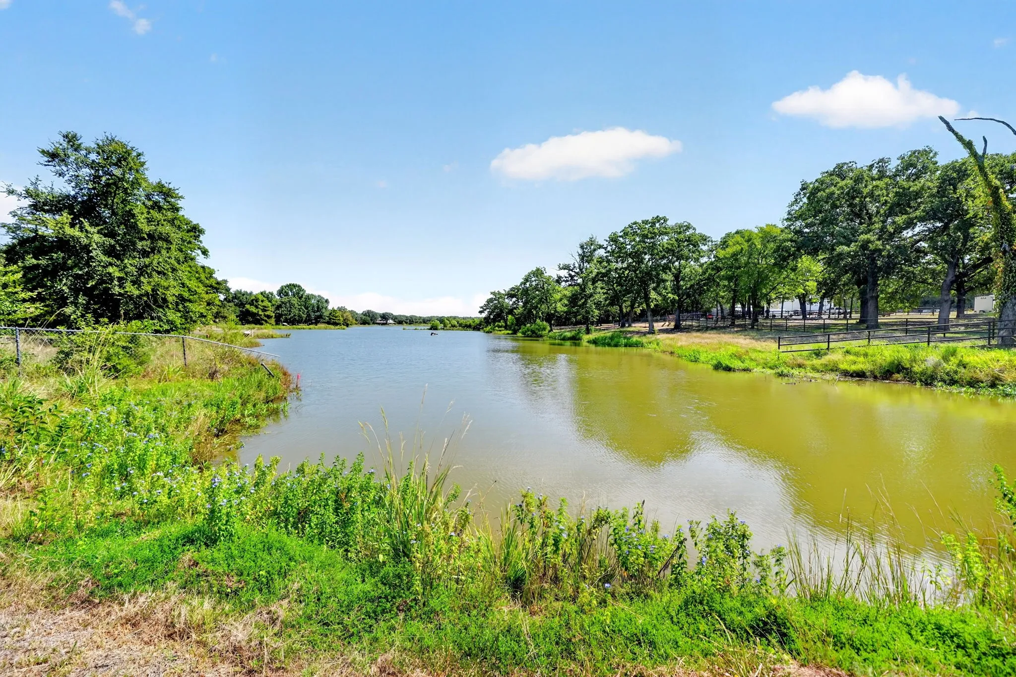 Expansive water feature with natural banks, surrounded by mature trees, and bordered by a wire fence
