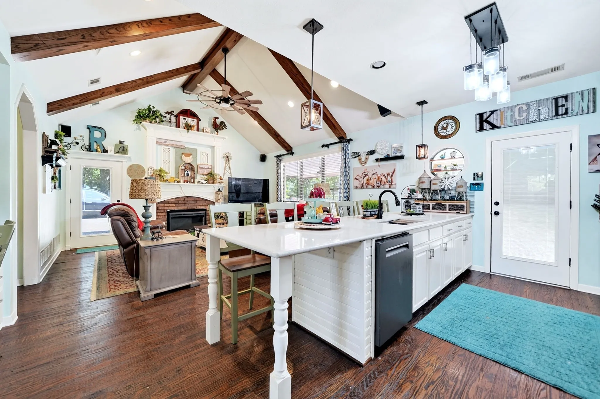 Expansive open-concept living area featuring a vaulted ceiling with exposed wood beams, wood-finish flooring, a brick fireplace with a white mantle, a central island with white cabinetry and a built-in dishwasher, and a glass-paned exterior door