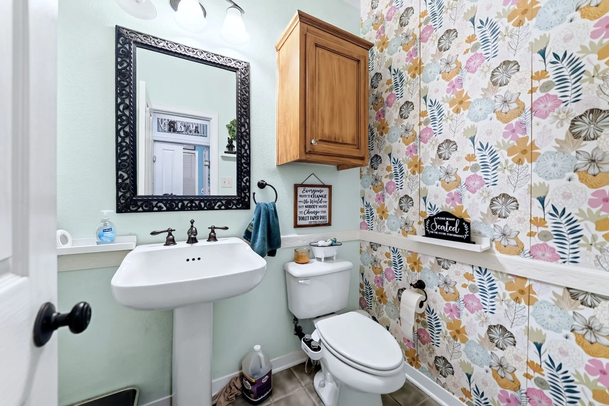 Pedestal sink with dark bronze faucet, framed mirror, floral wallpaper with wainscoting, and an overhead wooden cabinet