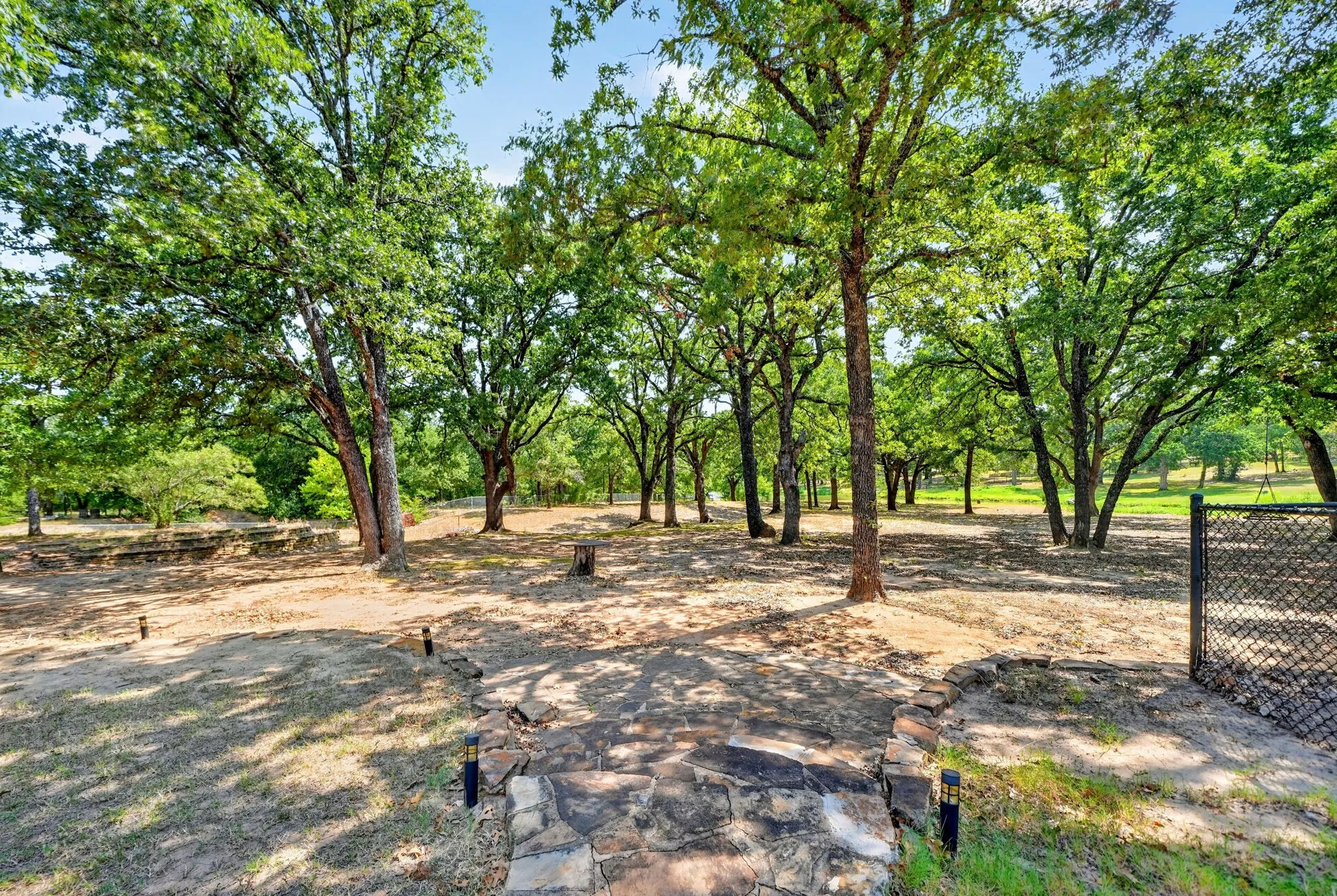 Expansive wooded grounds featuring numerous mature trees, a stone pathway, ground lighting fixtures, and a chain-link gate
