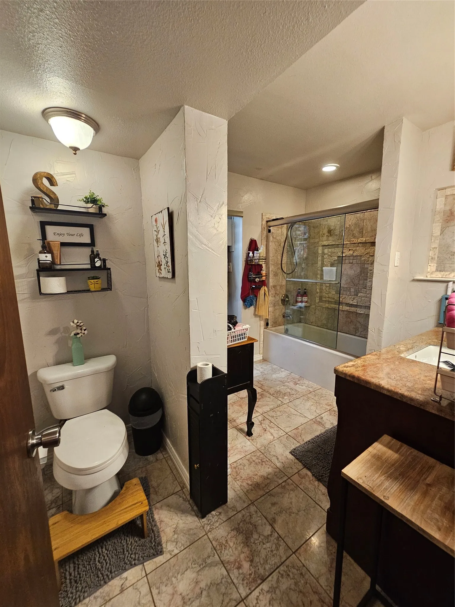 Bathroom featuring patterned floor tiling, a white toilet with wall-mounted shelving, a built-in bathtub with glass sliding doors, a vanity with a counter-mounted sink, and a sconce light fixture
