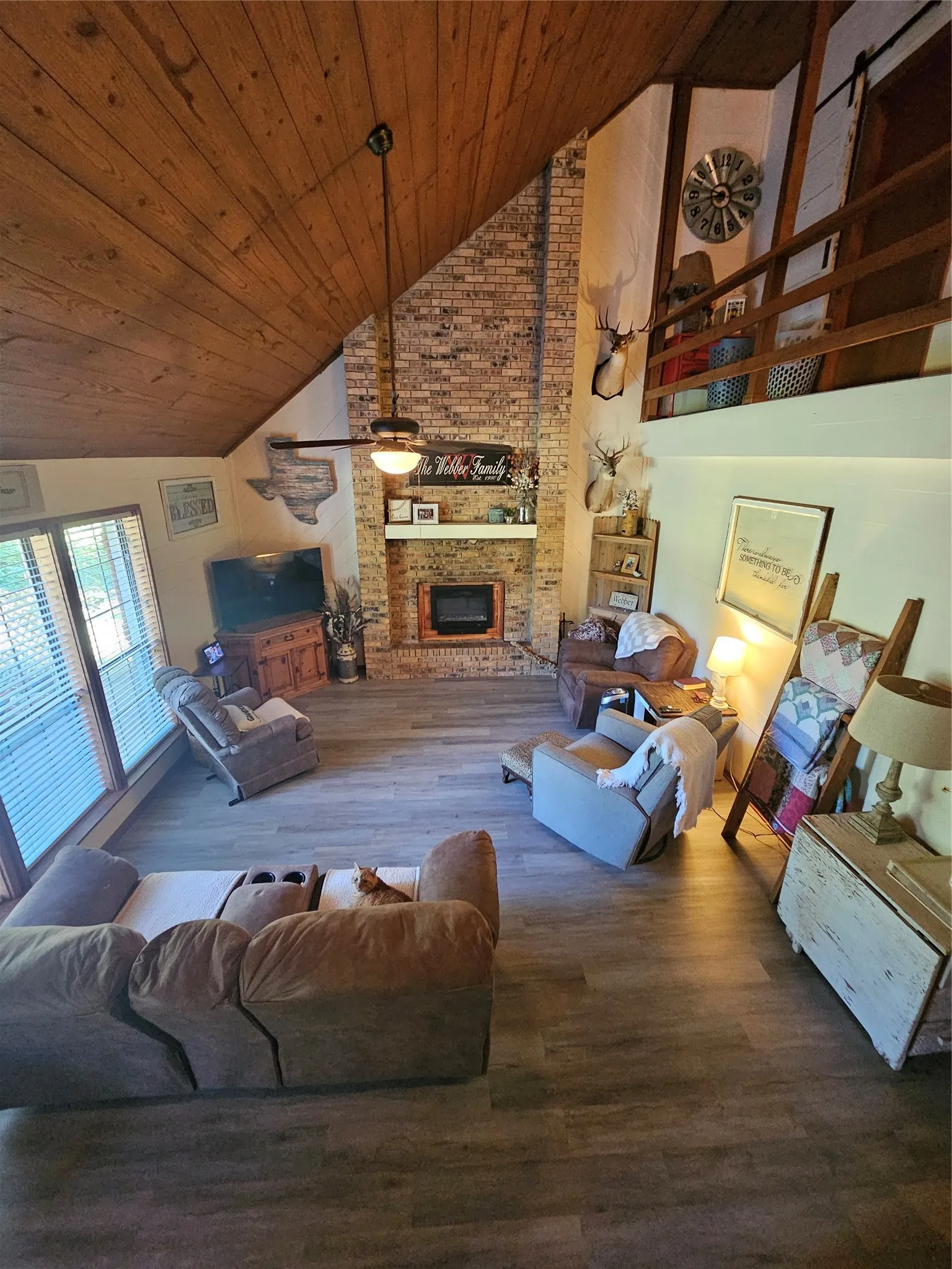 Spacious living area featuring a vaulted wood-paneled ceiling, a brick fireplace with a wood mantle, wood-finish flooring, and a large window with blinds