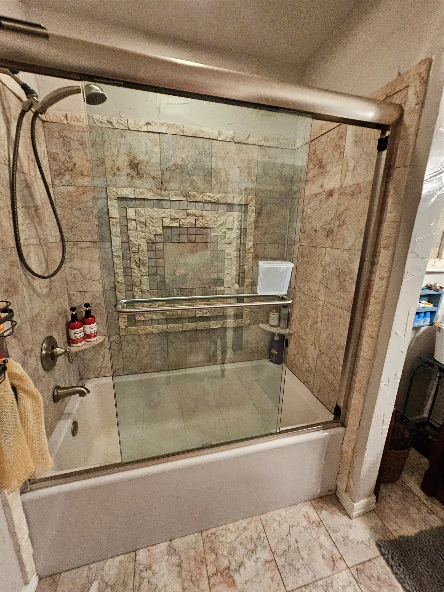 Bathtub with sliding glass shower door, brushed nickel hardware, and tile surround featuring a decorative mosaic inlay